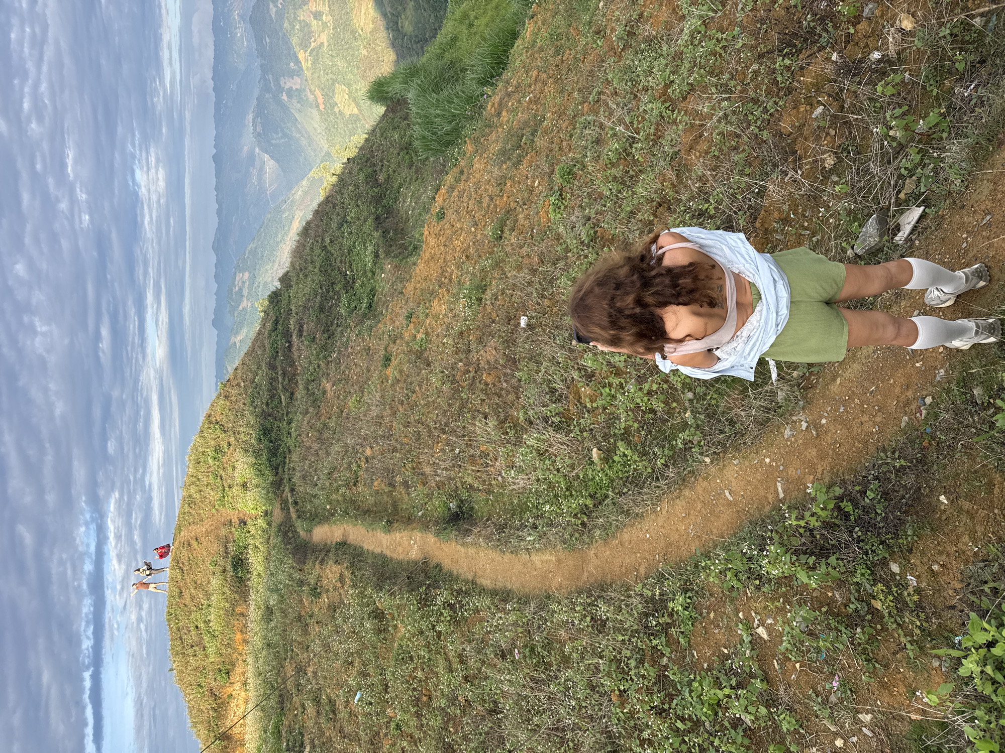 A high-angle shot depicts a landscape in Lũng Cú Commune, Vietnam. A woman with curly brown hair, wearing a light blue top, white bralette, green shorts, white socks, and grey sneakers, stands on a dirt path. The path winds through a grassy hillside towards a group of people gathered at the edge. In the distance, mountain ranges stretch under a cloudy sky.