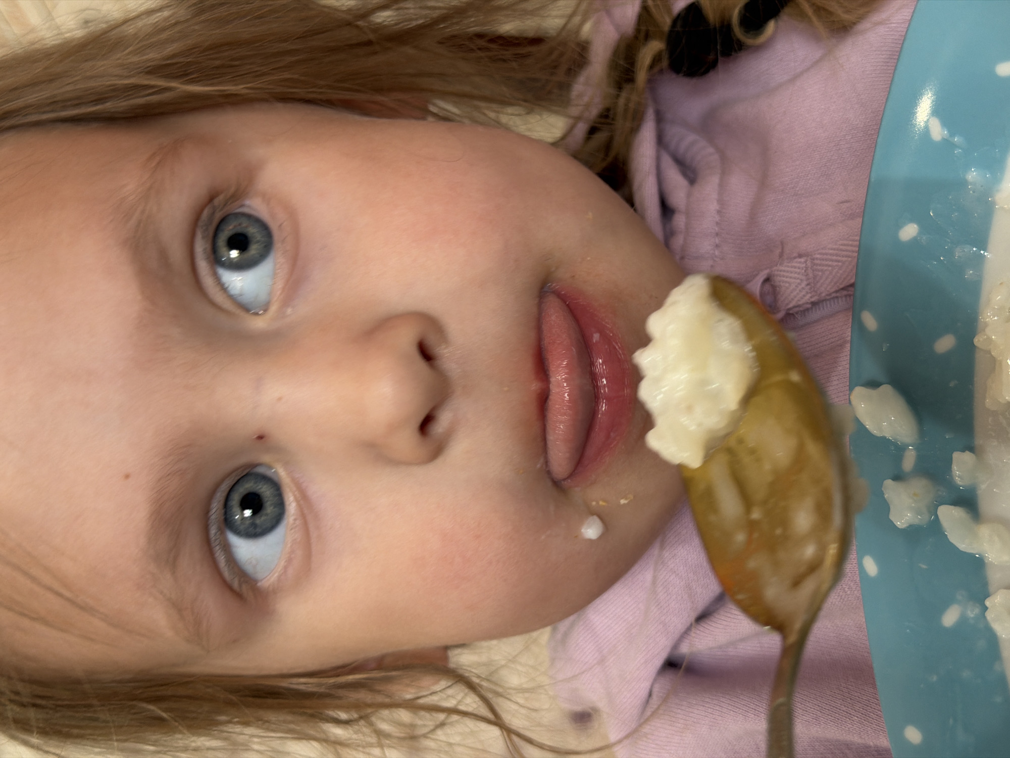 A young girl with fair skin and blonde hair is pictured close-up. She has blue eyes and is sticking her tongue out slightly. A spoonful of food, possibly rice pudding, is positioned near her mouth, and there is a small drop of the same food on her cheek. Part of a blue plate with white polka dots, also containing the food, is visible on the right. The girl wears a light purple shirt or sweatshirt.
The location of the scene in Chaikovsky, Russia is not discernible from the image provided.