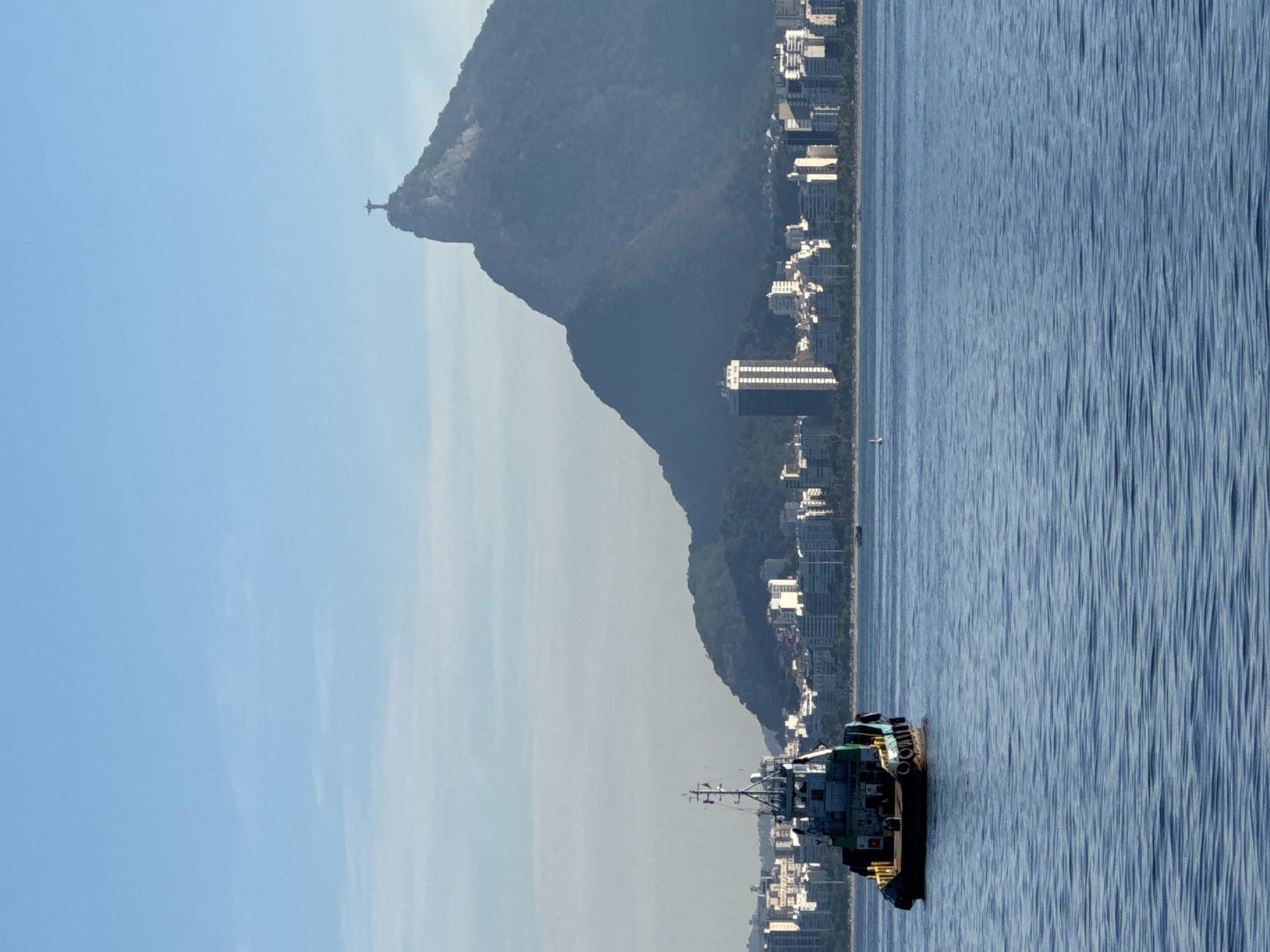 The image shows a tugboat in the foreground on the water, with a city skyline and a large mountain in the background. The tugboat is dark colored with yellow accents. The city skyline consists of numerous buildings, some of which are tall and modern, others appear to be residential buildings. The mountain behind the city is covered in lush green vegetation. Atop the mountain, a prominent statue of Christ the Redeemer is visible, overlooking the scene. The sky is a clear blue with a few faint clouds. The overall scene appears to be a daytime view of a coastal city. This is a view of Rio de Janeiro, Brazil, from the city of Niterói across Guanabara Bay.