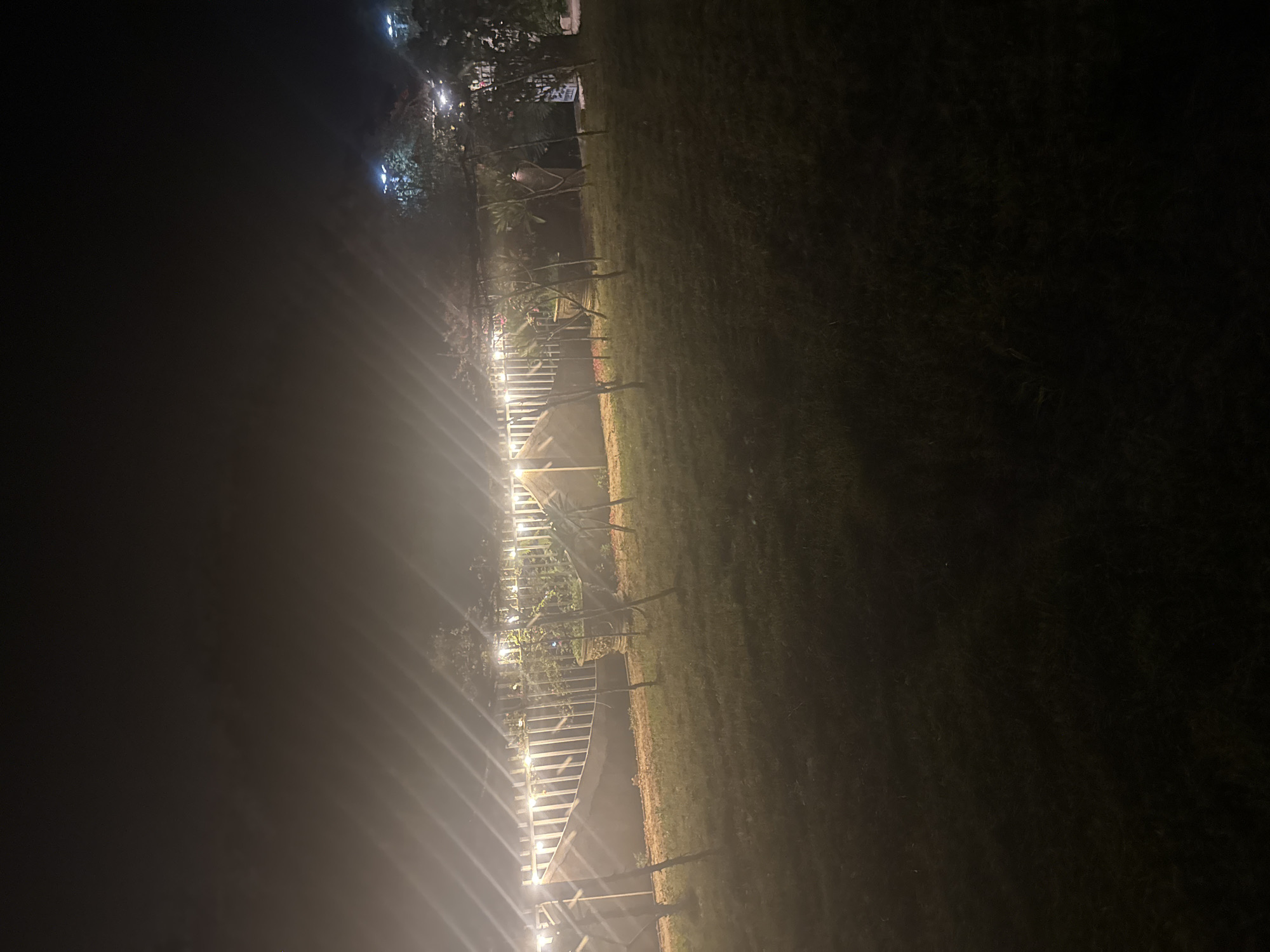 The image shows an outdoor scene at night, with a focus on a garden area illuminated by string lights. A low fence runs horizontally across the mid-ground, adorned with numerous small, bright string lights that cast a warm glow. Beyond the fence, trees and foliage are visible, some of which are lit by additional overhead lights, appearing as bright blue or white orbs. In the foreground, a grassy area is dimly lit, with several thin, vertical posts emerging from the ground, possibly part of a trellis or support structure for plants. A large, ornate planter is situated behind the fence, to the left of center, containing greenery. The overall atmosphere is somewhat hazy, suggesting fog or mist, which diffuses the light sources. No people or animals are clearly discernible in the image. The location is described as the city of Bac Nha Trang Ward, Vietnam.