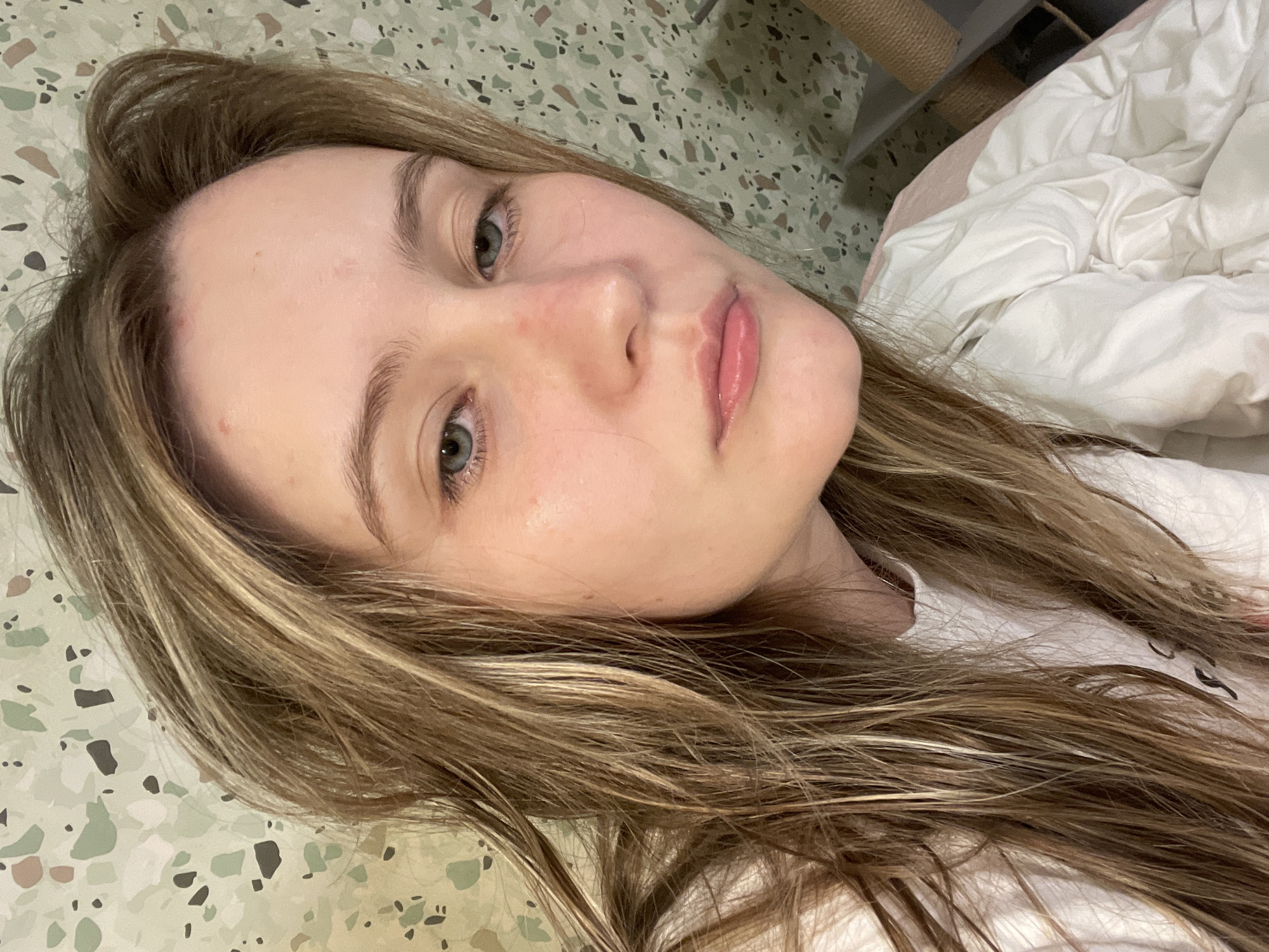 This is a close-up, low-angle photograph of a young person lying on their side, looking towards the camera. Their head is tilted, and their body is partially covered by a white duvet. The person has fair skin, light blue eyes, and long, wavy, light brown hair with blonde highlights. Their lips are slightly parted, and they appear to be wearing lip gloss. There are a few small reddish blemishes on their forehead and cheek.
The background is a patterned surface, possibly a floor or wall, with an abstract design of irregular shapes in shades of green, brown, and beige. To the right of the person, a white duvet with visible creases suggests a bed. A portion of a cat scratching post is visible in the upper right corner. The lighting appears soft and diffused, possibly indicating indoor lighting or natural light from a window. The overall mood is relaxed and intimate. There is no visible text in the image.