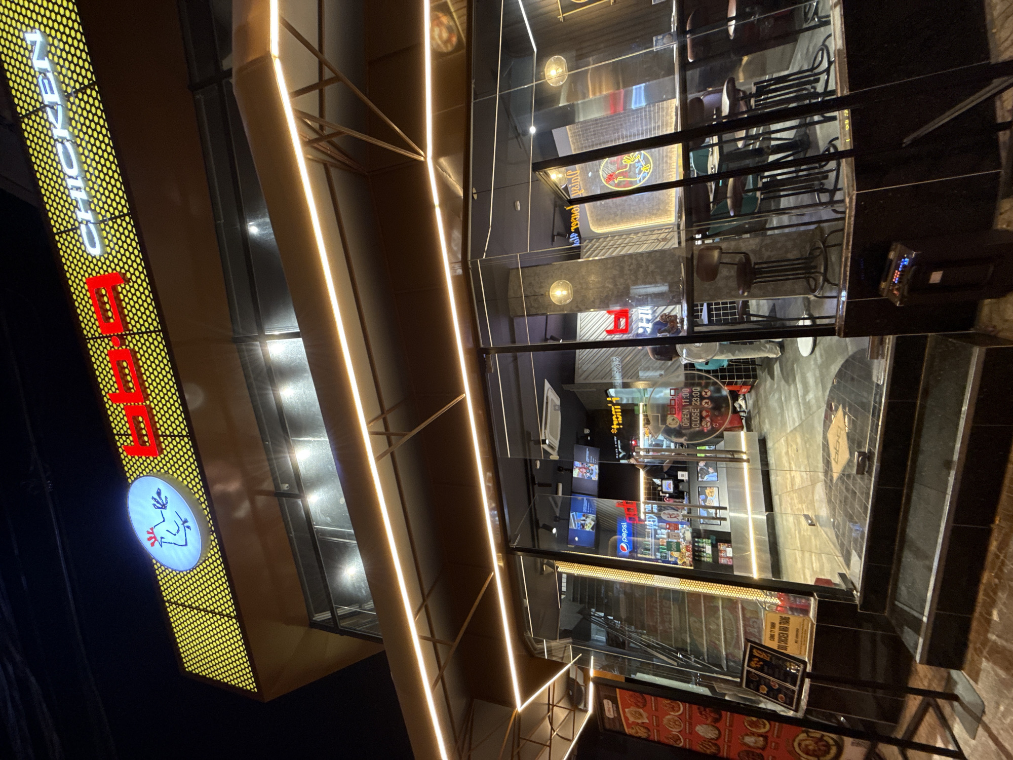 The image captures the exterior of a "BB.Q Chicken" restaurant in Nha Trang, Vietnam, at night. The establishment features a prominent yellow, dot-patterned facade with illuminated red and white signage displaying "BB.Q CHICKEN". A blue oval logo with a stylized white chicken is also visible on the signage. The entrance is marked by glass doors and a set of black tiled stairs leading up to the restaurant. Inside, a counter area is visible with shelves stocked with beverages, including Pepsi, and a display of food items. Seating arrangements with high stools and tables are present within the restaurant. A menu board is placed outside the entrance. The lighting is a mix of artificial lights from the signage, interior lights, and decorative string lights along the exterior. The overall scene depicts a modern commercial establishment in an urban setting.