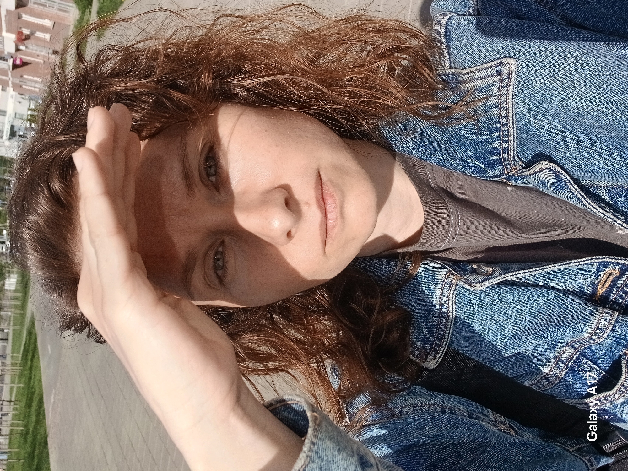 A fair-skinned woman with curly brown hair is shielding her eyes from the sun with her hand. She is wearing a denim jacket over a gray T-shirt. The photo appears to be taken outdoors on a paved surface, possibly a sidewalk or plaza. In the background, there are light-colored buildings. Based on the presence of "Galaxy A17" on the picture, this image may have been taken using a phone camera. It is not possible to determine the location of the photo as Yekaterinburg, Russia without additional information.