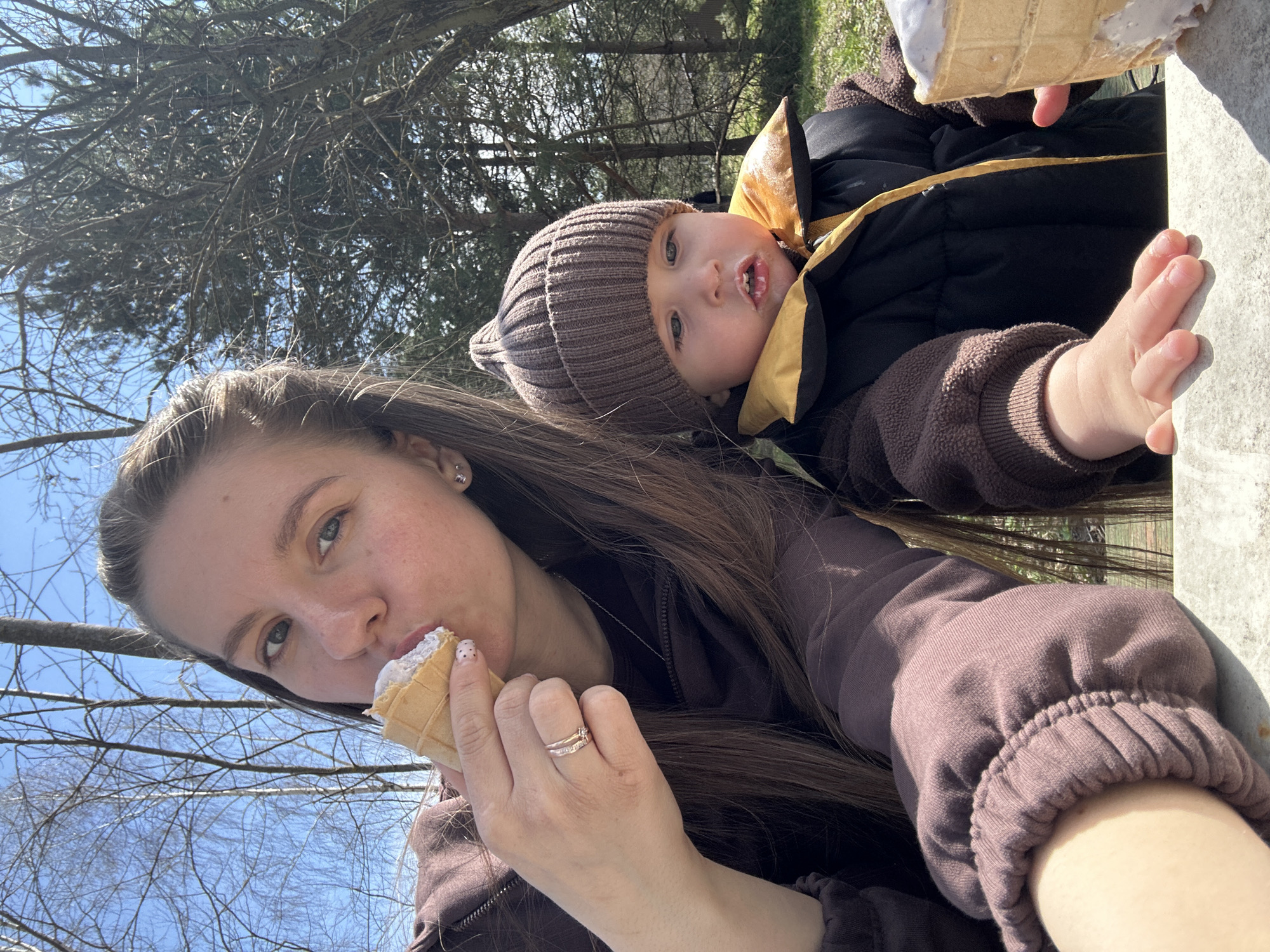 A woman and an infant are posing for a selfie outdoors. The woman has long brown hair and is wearing a brown hooded sweatshirt. She is holding an ice cream cone in her right hand, which she is partially eating. She is wearing a ring on her ring finger. An infant, dressed in a brown knitted cap, a yellow vest, and a black jacket, is being held in the frame to the right of the woman. The infant's hands are visible on the right side of the frame. There are trees and clear sky in the background. The location is outdoors, but no specific geographical landmarks are visible to determine the exact location within the city of Мещёры, Russia.