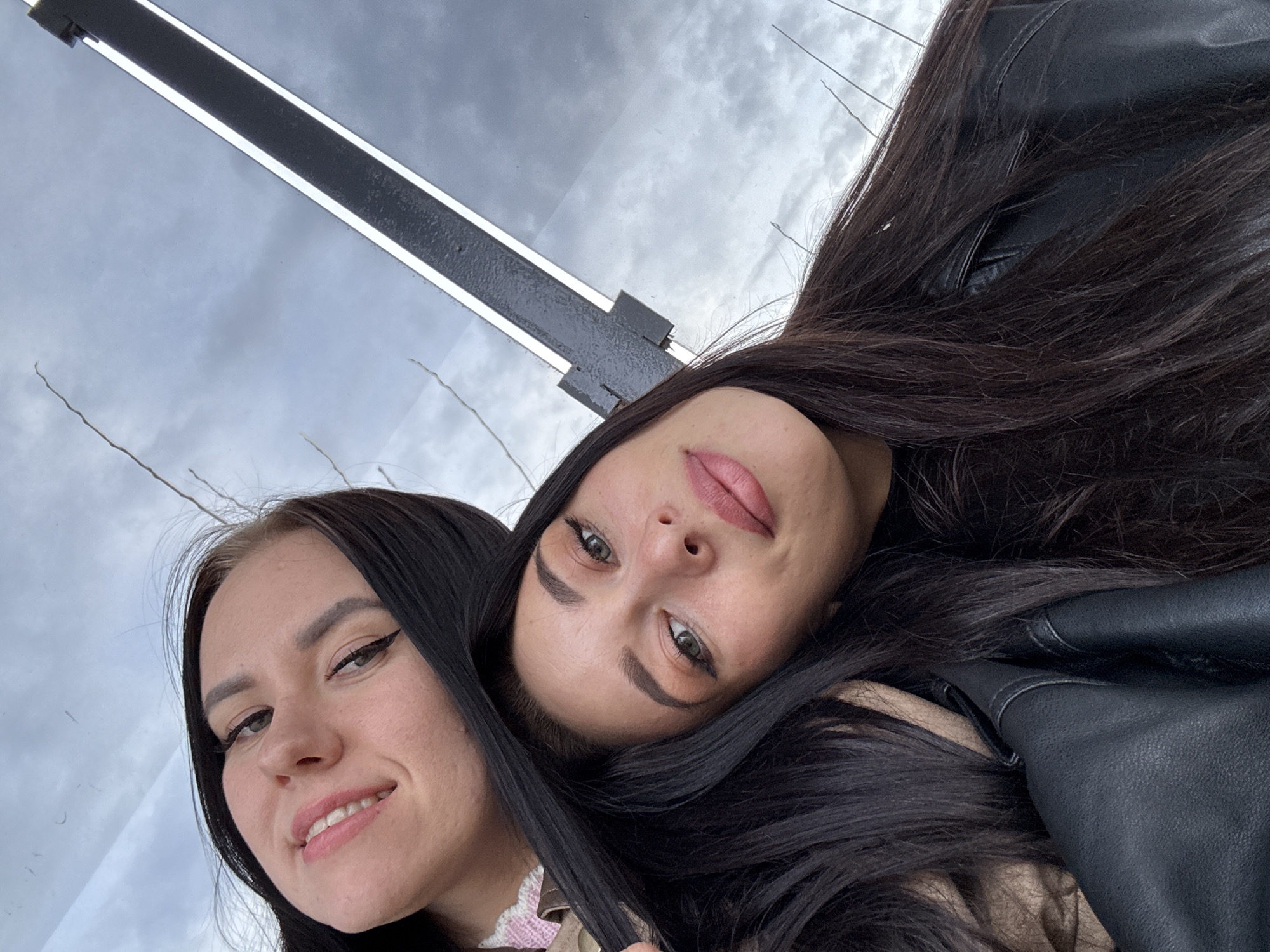 Two women pose for a selfie. The woman on the left has dark hair, fair skin, and is smiling with visible white teeth. Her eyes are enhanced with eyeliner. The woman on the right has dark hair and is angled slightly towards the camera. She has full lips, pronounced eyebrows, and light-colored eyes. She wears what appears to be a black leather jacket. The background includes a cloudy sky and a black metal pole with a white light fixture. The location is the city of Chaikovsky, Russia.