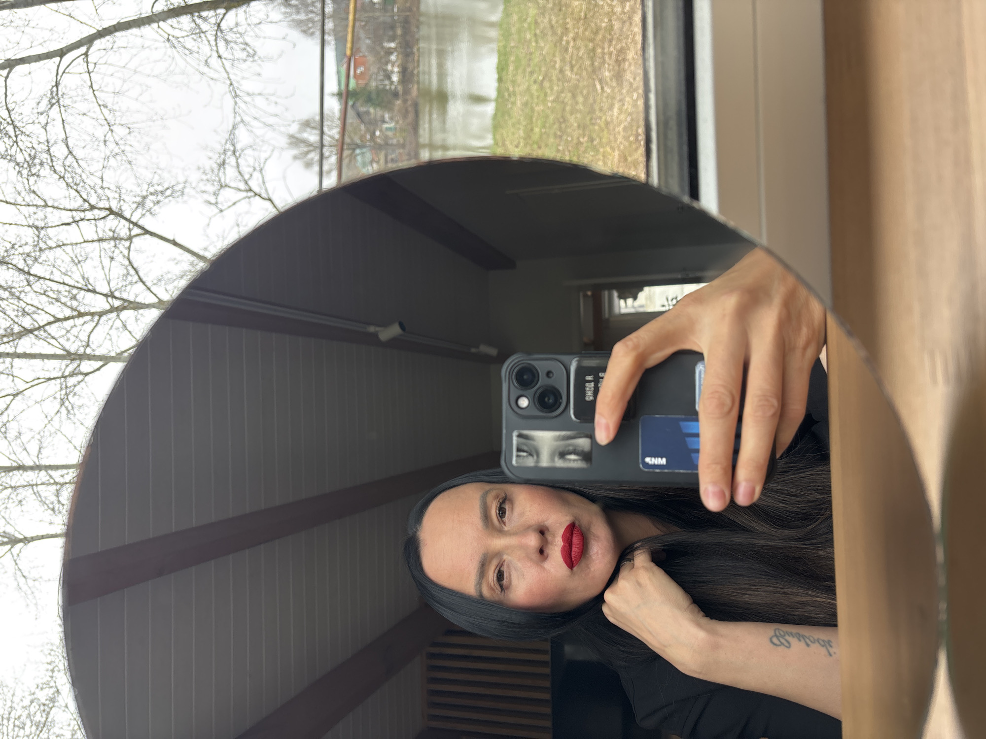 An adult woman takes a mirror selfie indoors, holding a dark-colored smartphone with a triple camera array in her right hand. She has long, dark hair, fair skin, and striking red lipstick, with a neutral expression as she looks directly into the camera. She wears a ring on her left ring finger and a dark top. A script tattoo, appearing upside down, is visible on her right forearm.
The smartphone case features a sticker with an image of eyes, and a blue card with the letters "SNM" is partially visible, tucked behind the phone. Additional upside-down text, possibly "RREA FR", is also visible on the phone case.
The large, rounded mirror reflects an interior with a wooden, slatted ceiling and exposed beams. A slatted wooden panel is visible behind the woman's head in the reflection. Through a window visible in the mirror's reflection, an outdoor scene is depicted: bare tree branches against a bright, overcast sky, a body of water (likely a river or lake) in the mid-ground, and yellowish-green grass on the bank. This outdoor setting, with its bare trees and body of water, is consistent with Chaikovsky, Russia, during late autumn, winter, or early spring. The overall impression is a casual, indoor setting with a scenic view outside.