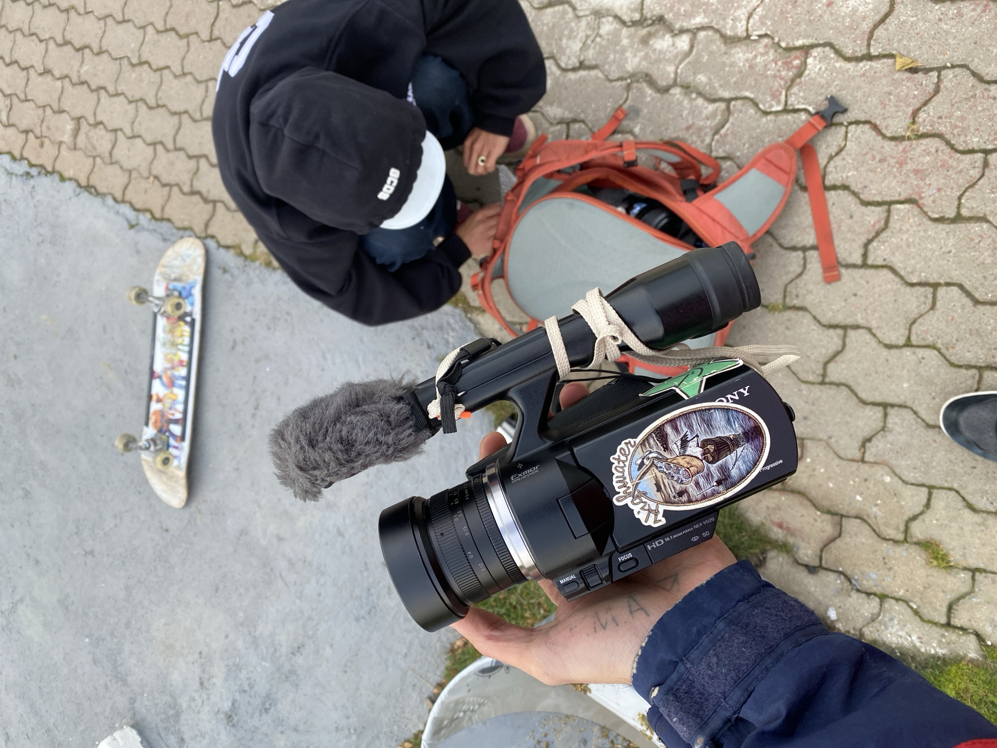 The image shows a street scene from an overhead perspective. A person in a black hooded sweatshirt and a cap is kneeling beside an open orange and gray backpack on a paved surface, possibly adjusting items within the backpack. To the left, there's a skateboard with colorful graphics lying on the ground.
In the foreground, a person is holding a Sony video camera with a microphone attached. The camera has a sticker on it featuring a bird and the word "Thwater". The person holding the camera is wearing a blue jacket with Velcro closures on the cuffs.
The pavement is light gray and appears to be made of interlocking stones, with some patches of moss or small vegetation growing in the cracks. Another person wearing a dark shoe is standing near the backpack and camera.
The location cannot be determined from this image. Ushuaia, Argentina is not determinable.