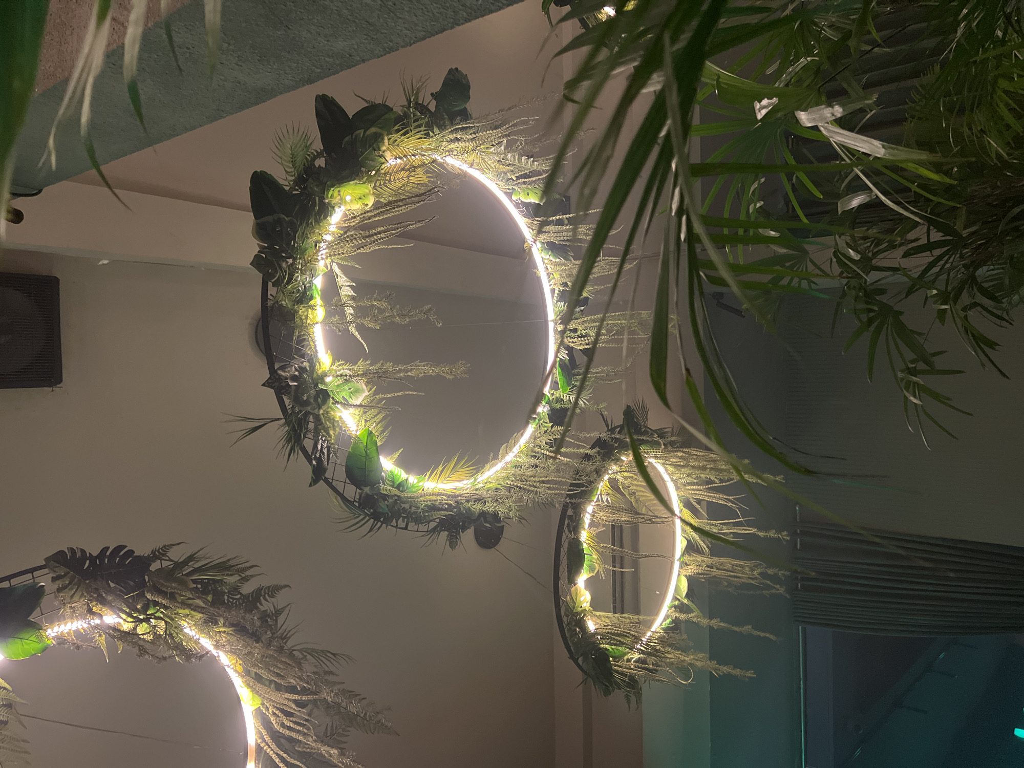 Captured from below, the scene features three illuminated circular decorations hanging from the ceiling, embellished with assorted green plants and fronds. A dark vent is visible on the upper-left, near the ceiling. A window can be observed on the bottom-right, with faint light emerging from it. Other plants with lengthy fronds are observed hanging in the top-right portion of the frame. The photograph appears to be taken in a space in North Kuta, Indonesia.
