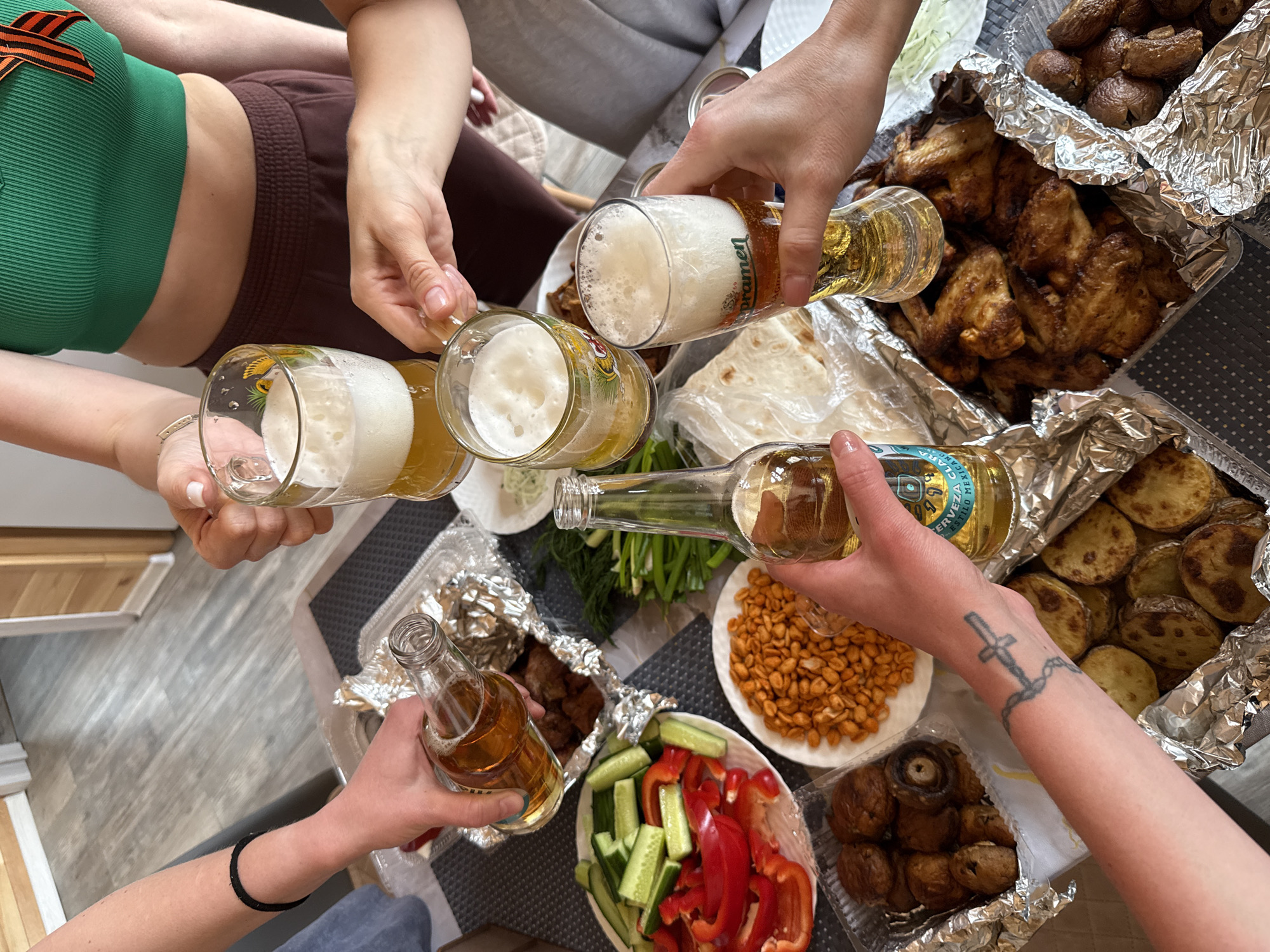 The scene shows a group of people toasting with beer and bottles, with a table covered in food. The food includes cooked chicken wings, potatoes, mushrooms, kebab-like meat, peanuts, sliced cucumbers and red bell peppers. In the top right, there is a bottle of "Pramen" beer. In other hands are Mexican beer bottles. One person is wearing a green top with a Saint George ribbon pinned to it. The scene is likely in Krasnoyarsk, Russia, due to the Russian-style food and the Saint George ribbon.