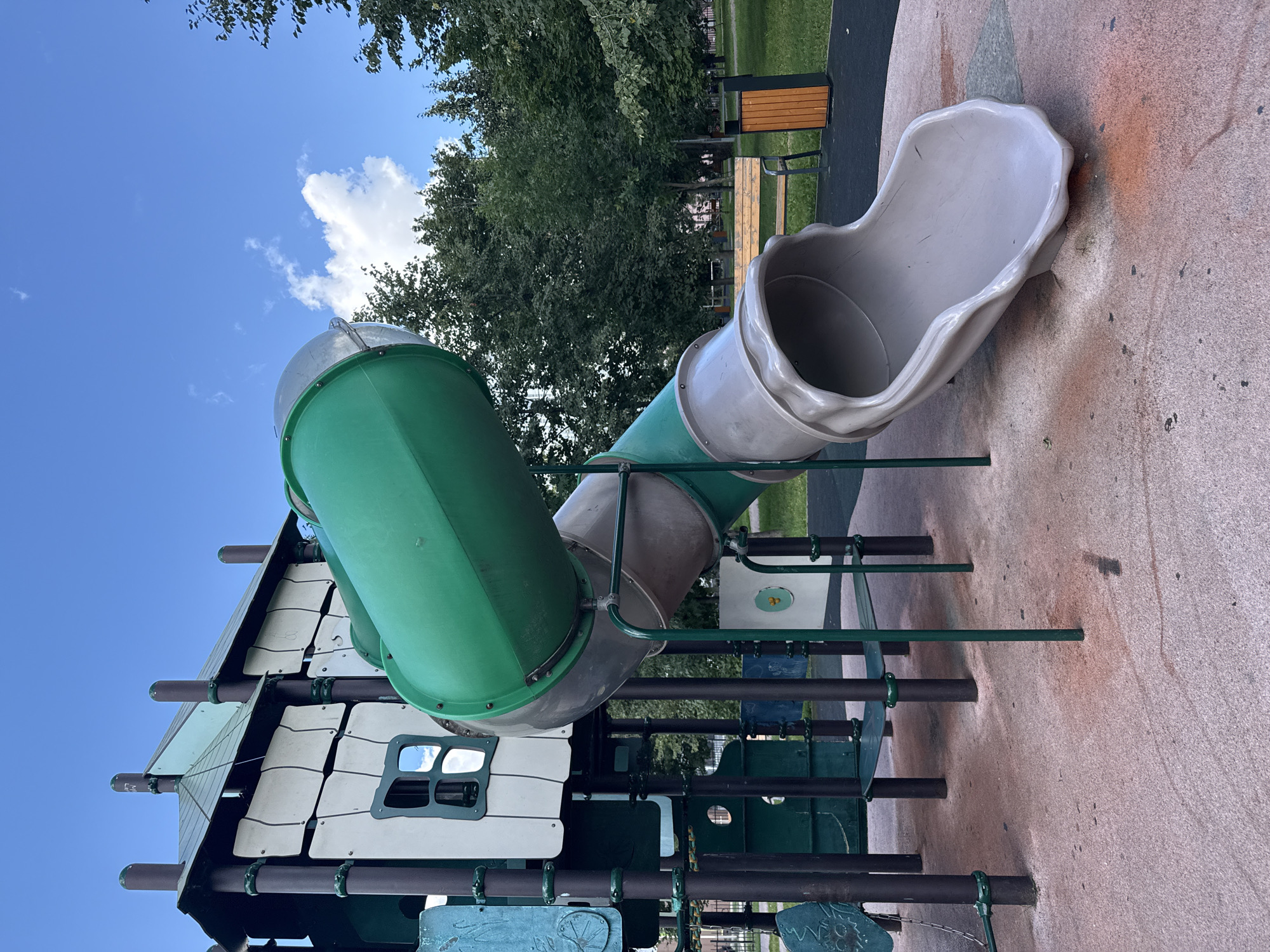 The image shows a playground slide in Lyubertsy, Russia. The slide is green and grey, with a tube-like structure. It is made of plastic and metal. The playground is located in a park with trees and grass. The sky is blue with some clouds. There is a bench and a trash can in the background. The ground is covered with a pinkish, speckled material. The playground structure has a house-like element with windows and a pitched roof.