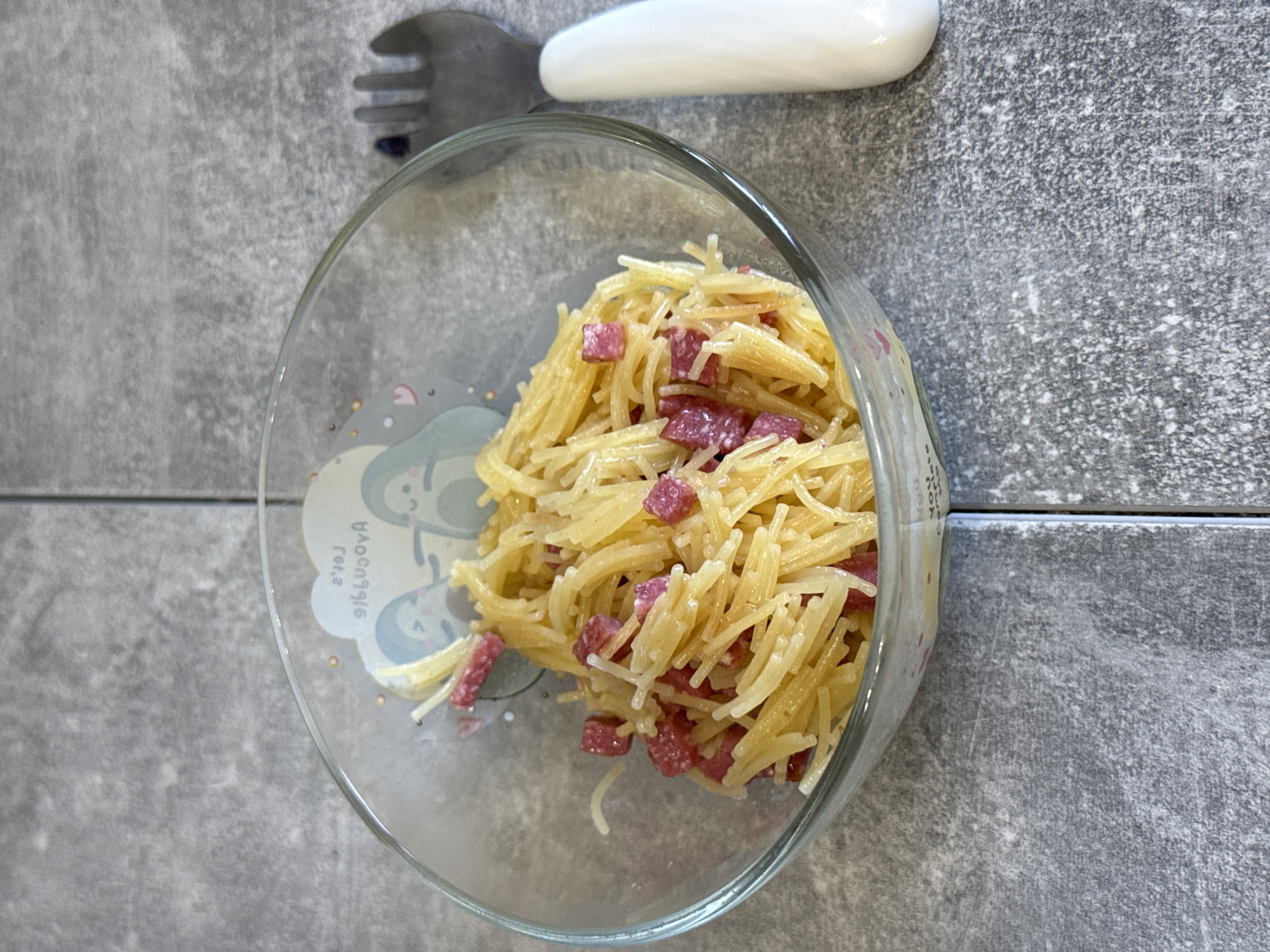 A close-up, eye-level shot shows a glass bowl filled with thin spaghetti mixed with small cubes of red salami, placed on a grey, textured surface that resembles a tiled countertop. To the right of the bowl, a white-handled fork with dark tines is partially visible. The bowl itself has a decorative border featuring cartoon avocados. The background is a continuation of the grey, textured surface. The scene is devoid of any people or animals.