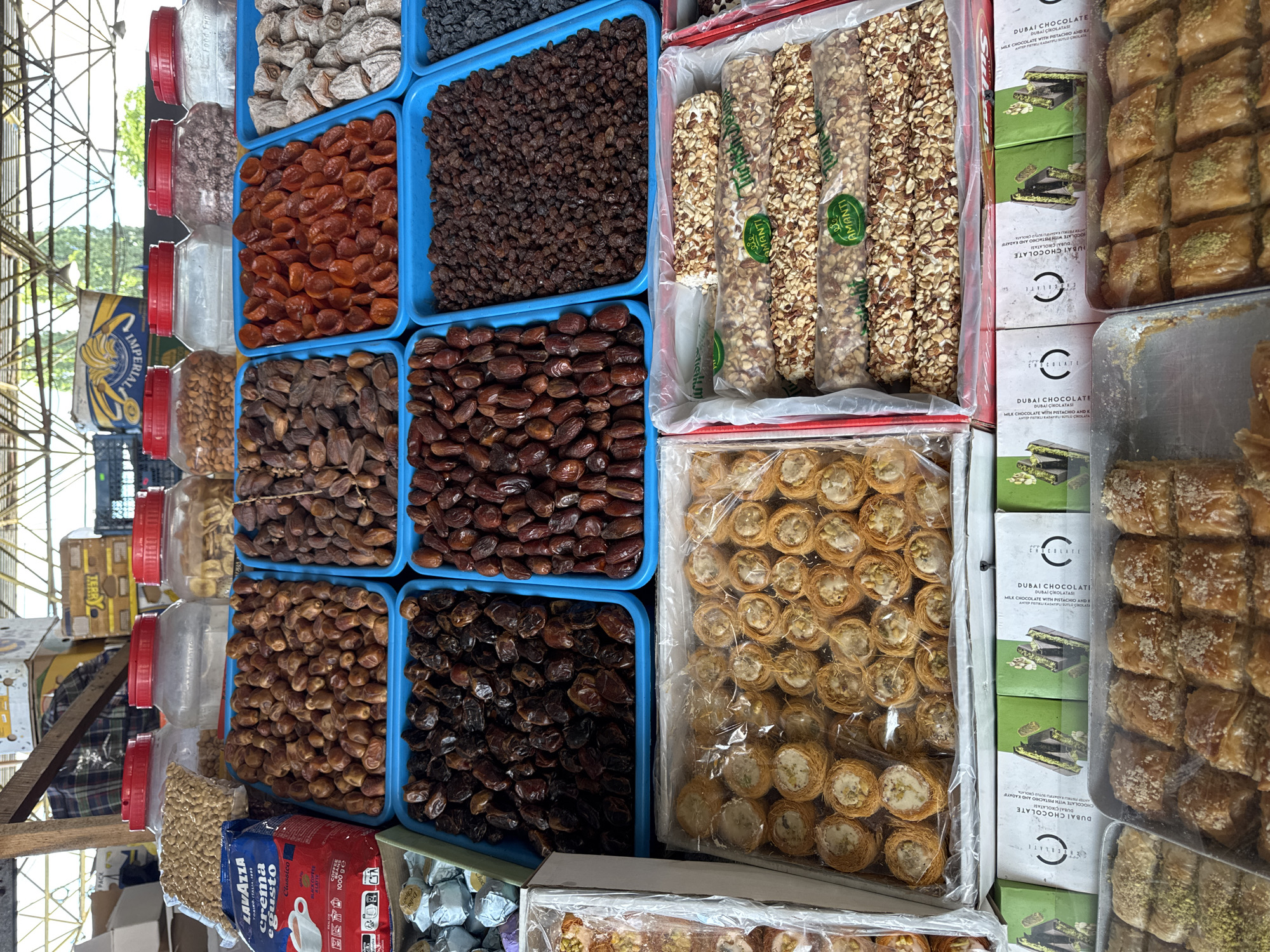 This image captures a vibrant display of dried fruits, nuts, and pastries at an outdoor market, likely in Odesa, Ukraine. The stall is laden with an array of goods organized in blue plastic trays and clear containers. Prominently featured are large quantities of dates in various shades of brown, as well as dried apricots and raisins. Several plastic jars with red lids hold an assortment of nuts and possibly other dried fruits. In the foreground, trays of sweet pastries, including what appear to be baklava and small, rolled confections filled with nuts, are displayed. Boxes of "Dubai Chocolate" are also visible, suggesting a diverse range of confectionery products. The market setting is suggested by the overhead structure with metal beams and translucent roofing, allowing natural light to illuminate the scene. The abundance and variety of goods indicate a bustling marketplace.