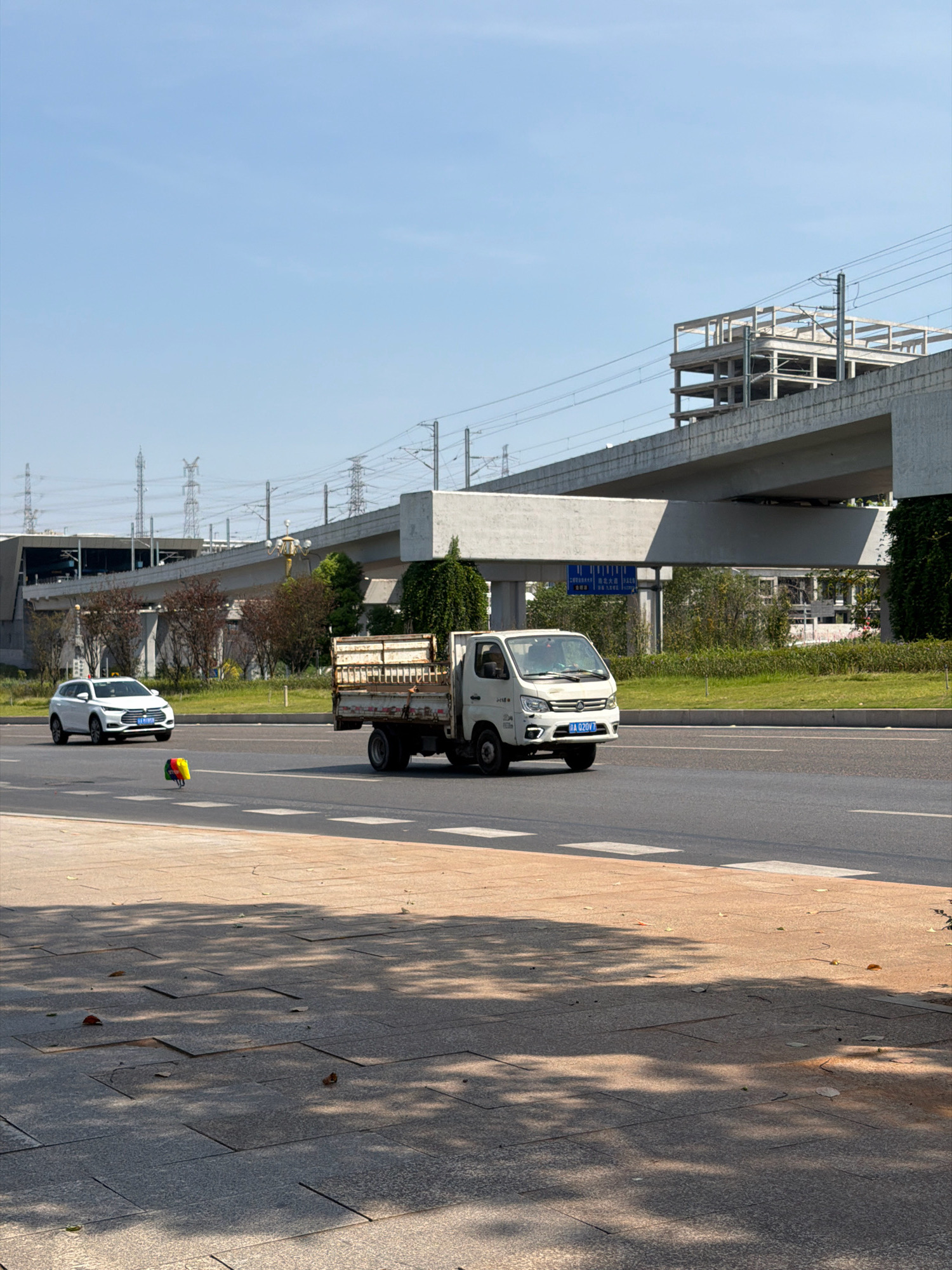 An outdoor, daytime, medium shot captures a street scene in Jiangjin, China. The focus is on a white, four-door vehicle, possibly a sedan or SUV, traveling on the right side of the road. To its right, a white pickup truck with an open bed is also moving along the same road. In the foreground, the ground is paved with large, rectangular stone tiles, dappled with sunlight and shadow. The middle ground shows the paved road with lane markings, and the background reveals an elevated structure, likely a bridge or overpass, under construction or recently completed, with visible concrete beams and supports. Beyond the elevated structure, power lines and transmission towers are visible against a clear blue sky. A small, colorful object, possibly a balloon or toy, rests on the road between the two vehicles.