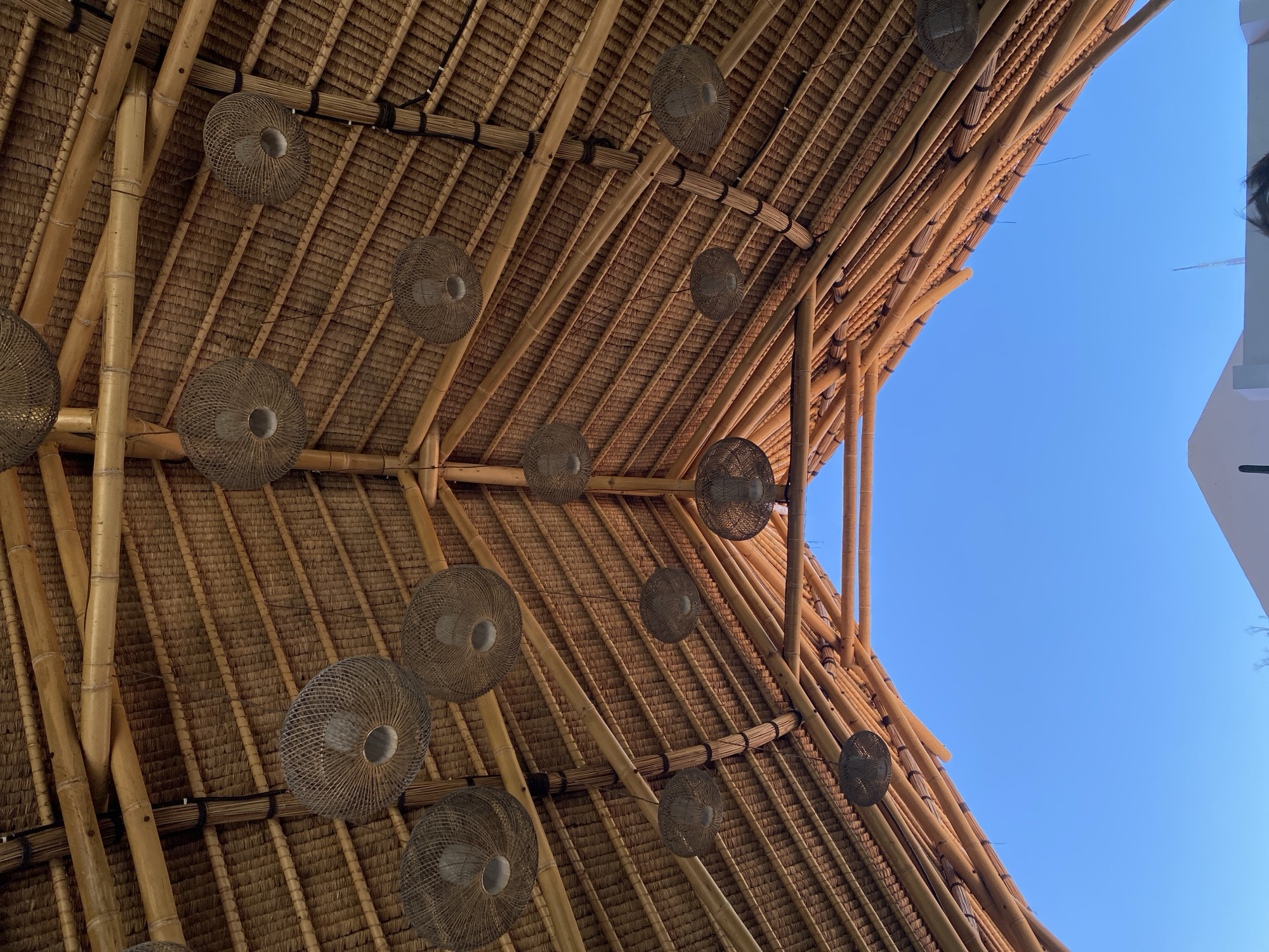 An upward-angled, outdoor shot shows the interior of a bamboo structure under a clear blue sky. The structure's roof is constructed of bamboo poles and thatched material, adorned with several round, woven light fixtures hanging from the beams. A portion of a white building with dark hair in the upper right corner peeks into the frame next to the bamboo. The photograph was taken at the city of Kuta Selatan, Indonesia.