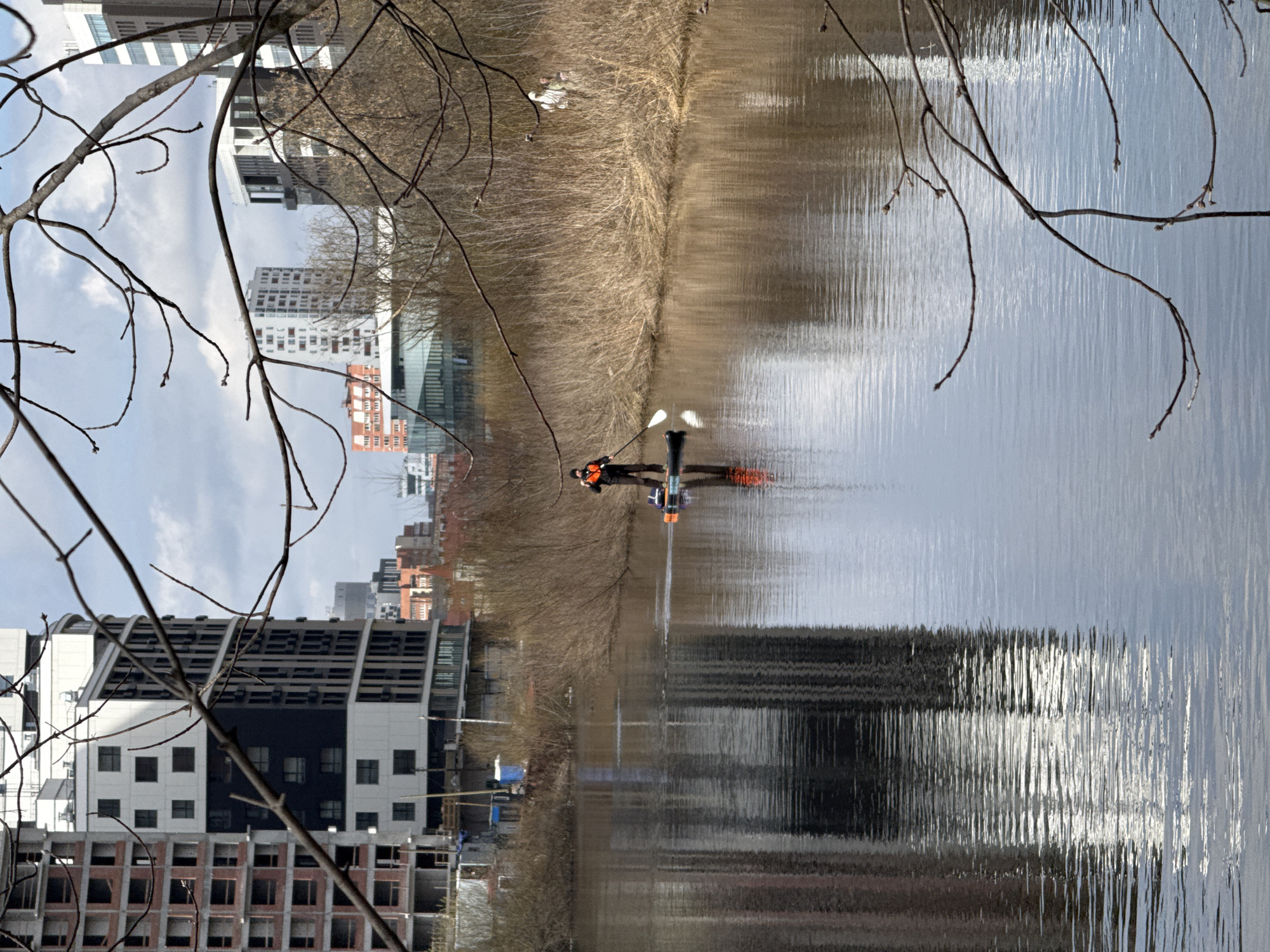 The image shows a person stand-up paddleboarding on a body of water, likely a river or canal, with urban buildings visible on the banks. The city is Yekaterinburg, Russia. The person is wearing a dark jacket and an orange life vest. They stand on a paddleboard with orange fins and hold a paddle. Bare branches frame the view in the foreground. The water reflects the sky, buildings, and trees on the opposite bank. The buildings range from modern high-rises to older, lower-rise structures. The riverbank is lined with trees lacking leaves. The sky is overcast.