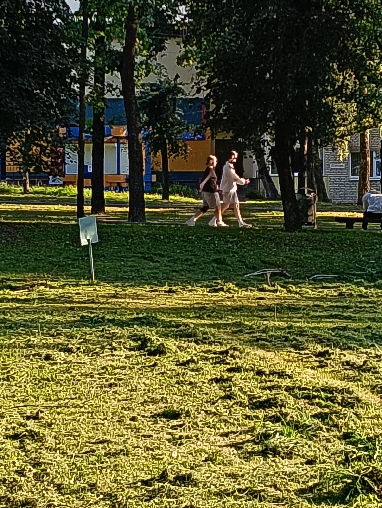 The image shows a grassy area with a few trees and buildings in the background. Two people are walking on the grass, and another person is sitting on a bench. A signpost is visible in the foreground. The scene appears to be a park or public space, possibly in Hrodna, Belarus, given the architectural style of the buildings and the overall atmosphere. The lighting suggests it is daytime, with sunlight casting shadows on the grass. The grass itself is a mix of green and yellowish hues, with some areas appearing freshly cut.