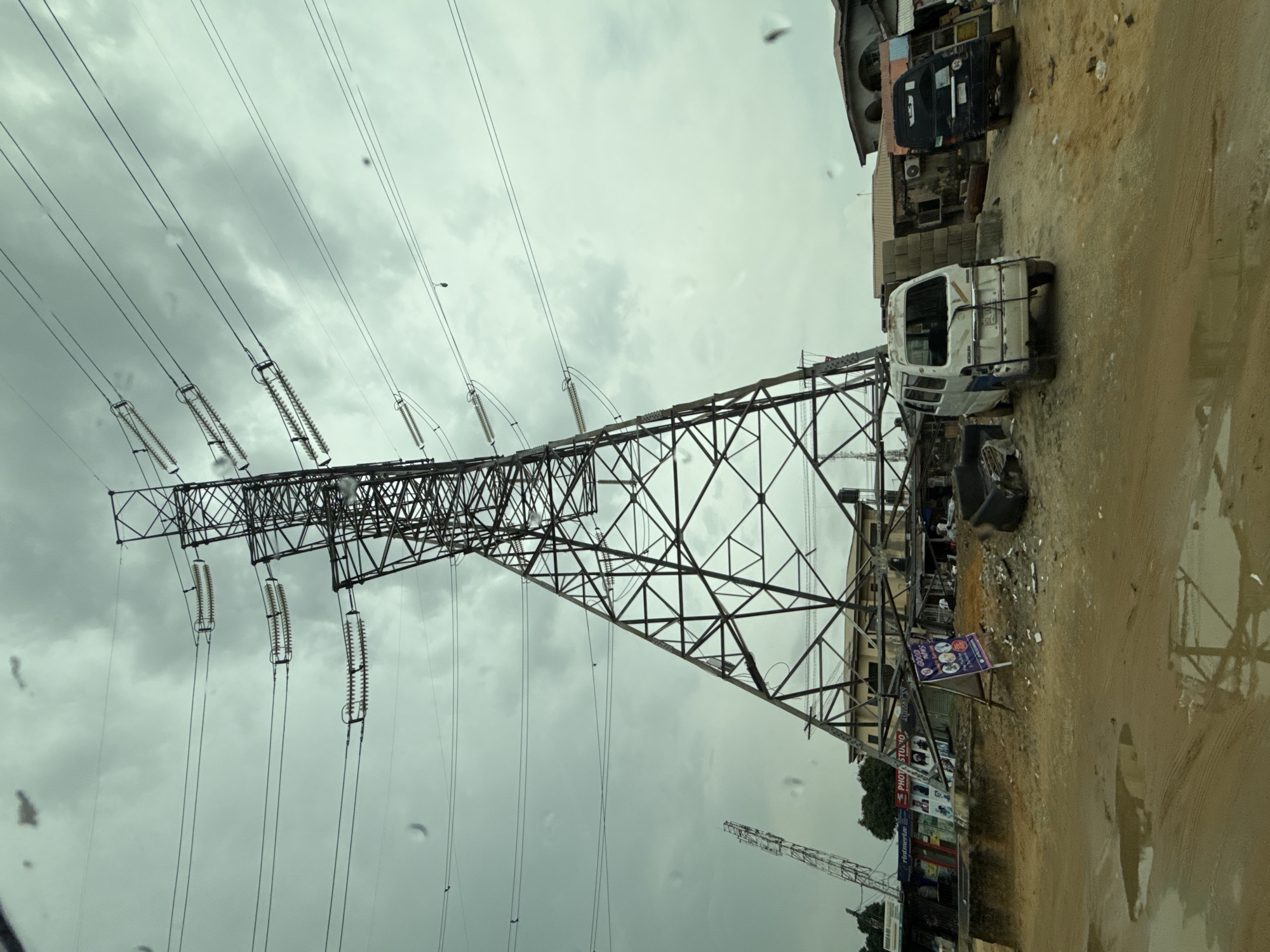 The image depicts a high-voltage electricity pylon in Lagos, Nigeria, under an overcast sky. The pylon, a large steel lattice structure, supports multiple high-tension wires. Adjacent to the pylon is a dirt road with puddles, and alongside the road are buildings of varying conditions. Discarded vehicles and construction materials are strewn about, adding to the disheveled urban environment. A photo studio, identifiable by signage, is visible amidst the surrounding structures.