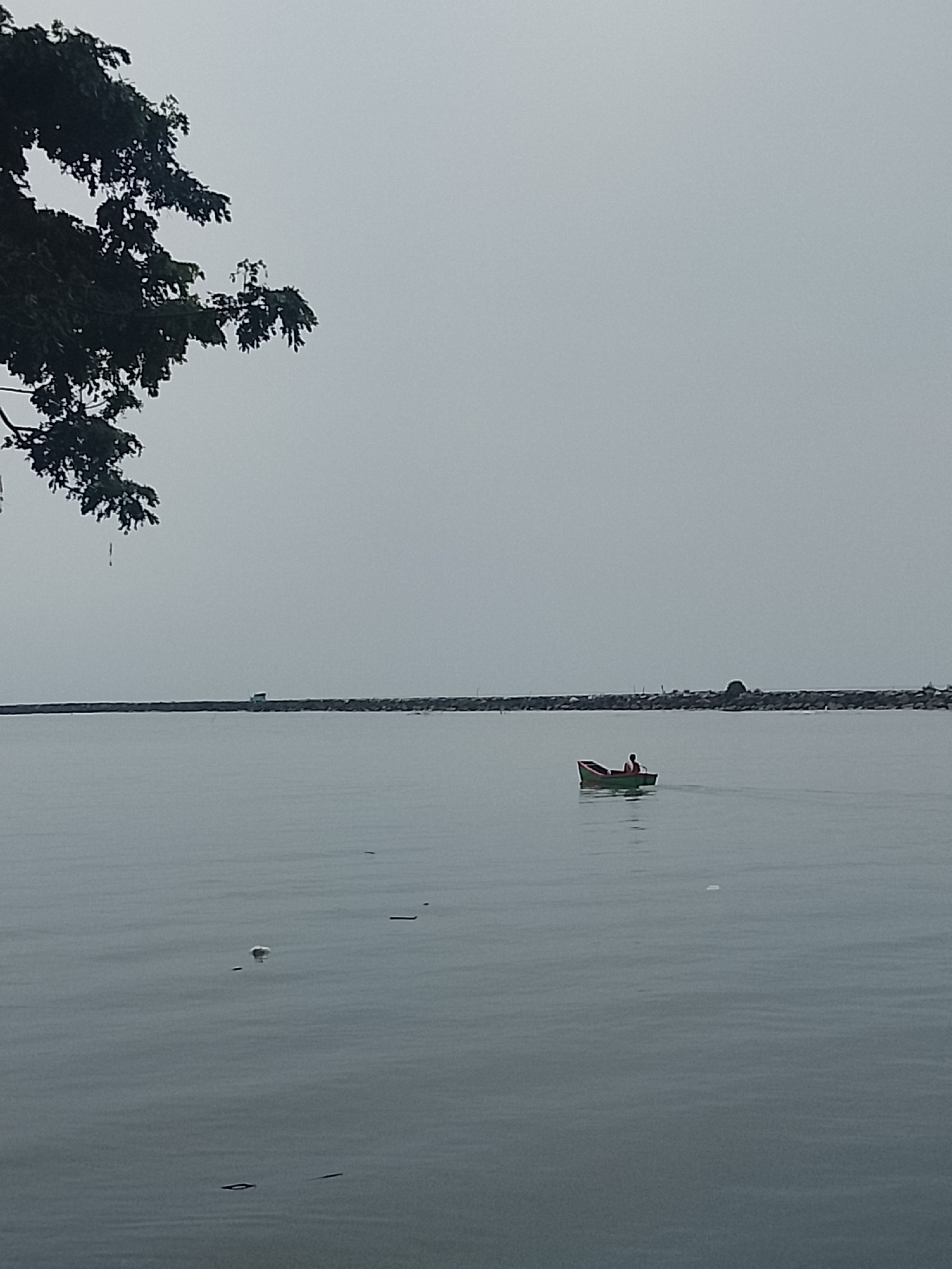 A person is in a small boat in the water. The boat is green and red. The person is wearing a red shirt and dark pants. The person is facing away from the camera and appears to be rowing. In the background, there is a long stone breakwater. The sky is overcast. The image appears to be taken from the shore of Reudeuep, Indonesia.