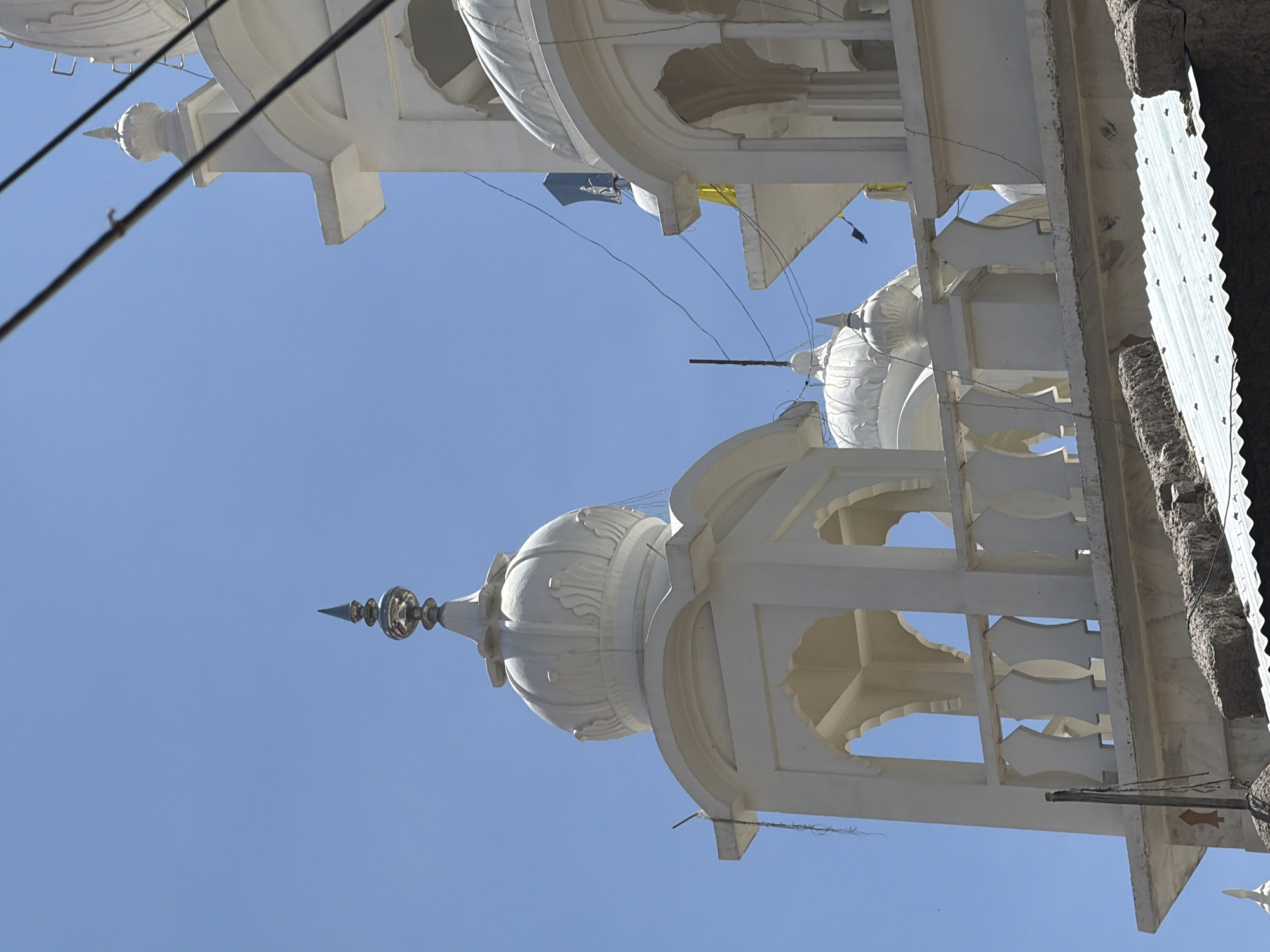The image shows the exterior of a white building with multiple domes and arches, set against a clear blue sky. The architecture suggests a religious structure, possibly a Gurdwara, given its ornate domes and spire-like finials. The building's details include decorative railings along a balcony or walkway. Power lines crisscross the frame, partially obscuring the view of the structure. In the foreground, a corrugated metal roof is visible, with some rough-textured building materials resting on it, indicating a more rustic or aged section of the site. The overall impression is of a bright, sunny day in an urban or semi-urban environment. The question of whether this is in Leh, India, cannot be answered from the image alone, as the image lacks specific landmarks or contextual cues to identify its location.