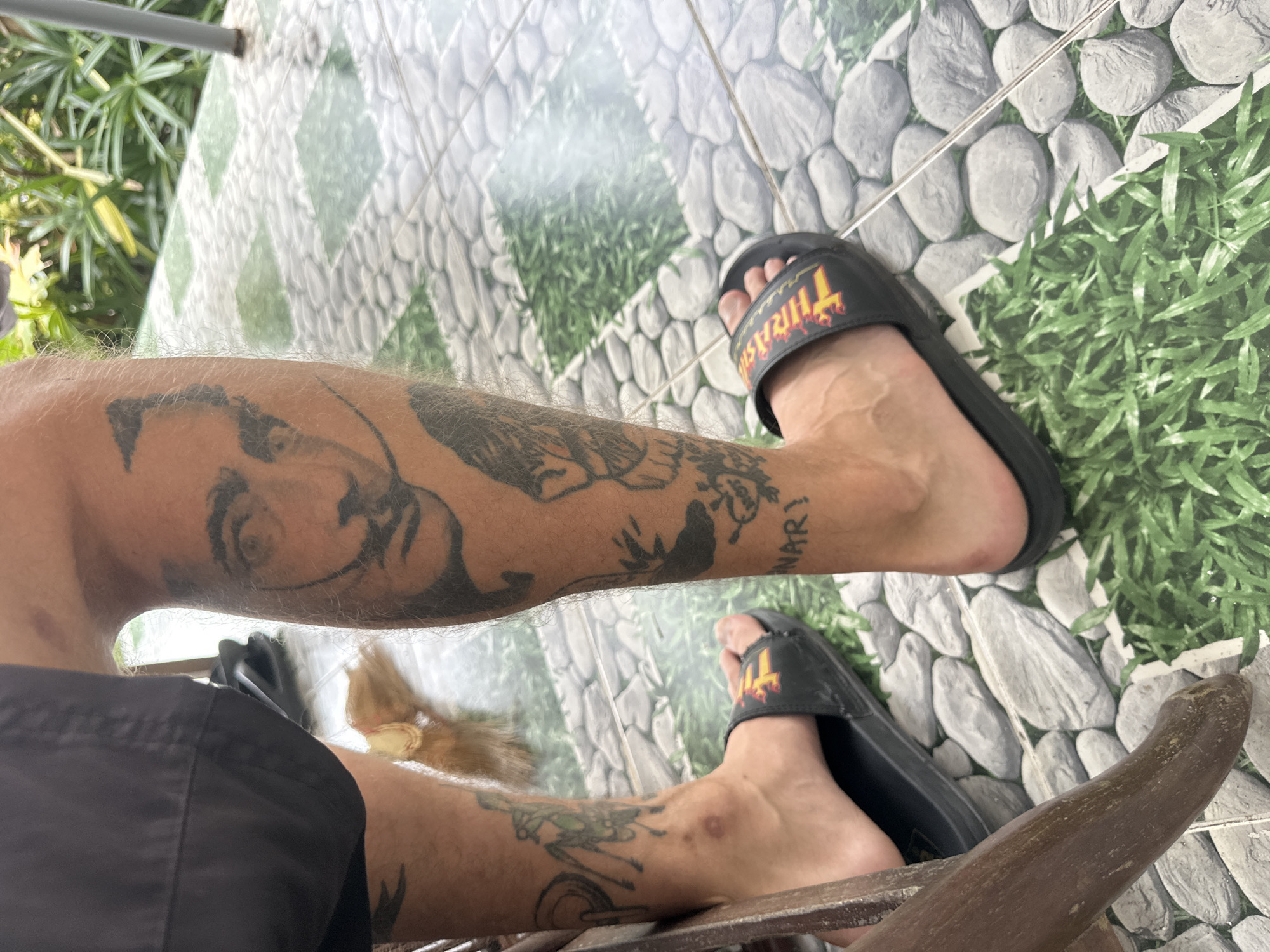 A person's legs are shown from the knees down, standing on a tiled patio. The tiles depict a pattern of grey stones and green grass. The person is wearing black shorts and black slide sandals with a red and yellow logo. Tattoos are visible on both legs. The right leg features a large tattoo of a face, resembling Salvador Dali. Below this, there is a tattoo of a skull and some text that reads "JNARY!". The left leg has a tattoo of an insect-like creature. A brown wooden chair leg is partially visible in the foreground. The background shows lush green foliage, suggesting an outdoor setting, possibly Karon, Thailand.