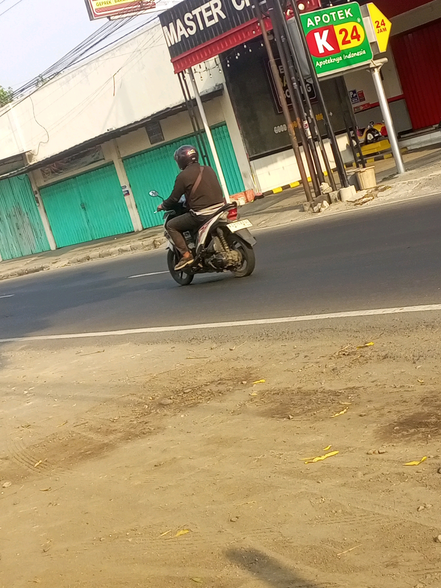 A person wearing a black long-sleeved shirt, dark pants, and a black helmet is riding a white and red motorcycle on a paved road in Gurah, Indonesia. The motorcycle has a white license plate. In the background, there is a building with green roll-up doors and a sign that says "MASTER C". To the right of "MASTER C" is a sign for "APOTEK K 24" which indicates it is a pharmacy open 24 hours. There are also several utility poles and wires. The ground in the foreground is a dirt and gravel surface with some yellow debris scattered on it. The lighting suggests it is daytime.