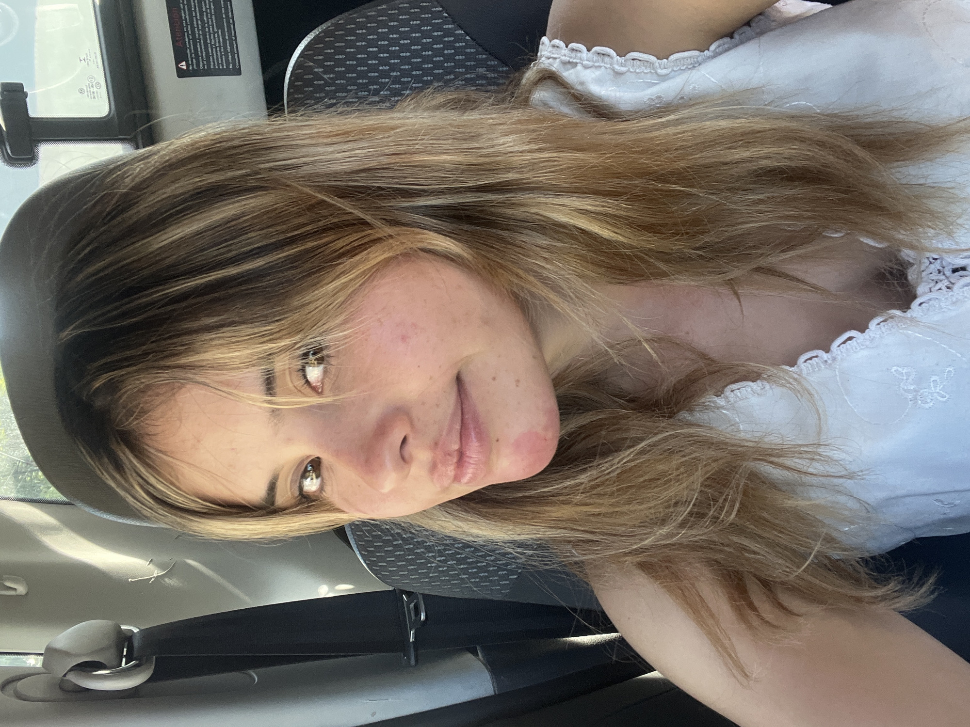 A young woman with long, light brown hair and fair skin is in a vehicle, likely a car, with a seat belt fastened. She is wearing a white top with lace trim. The interior of the vehicle is visible, including the seat and dashboard. The location is likely in a city, but not explicitly San Fernando del Valle de Catamarca, Argentina. No specific elements confirm the geographic location.