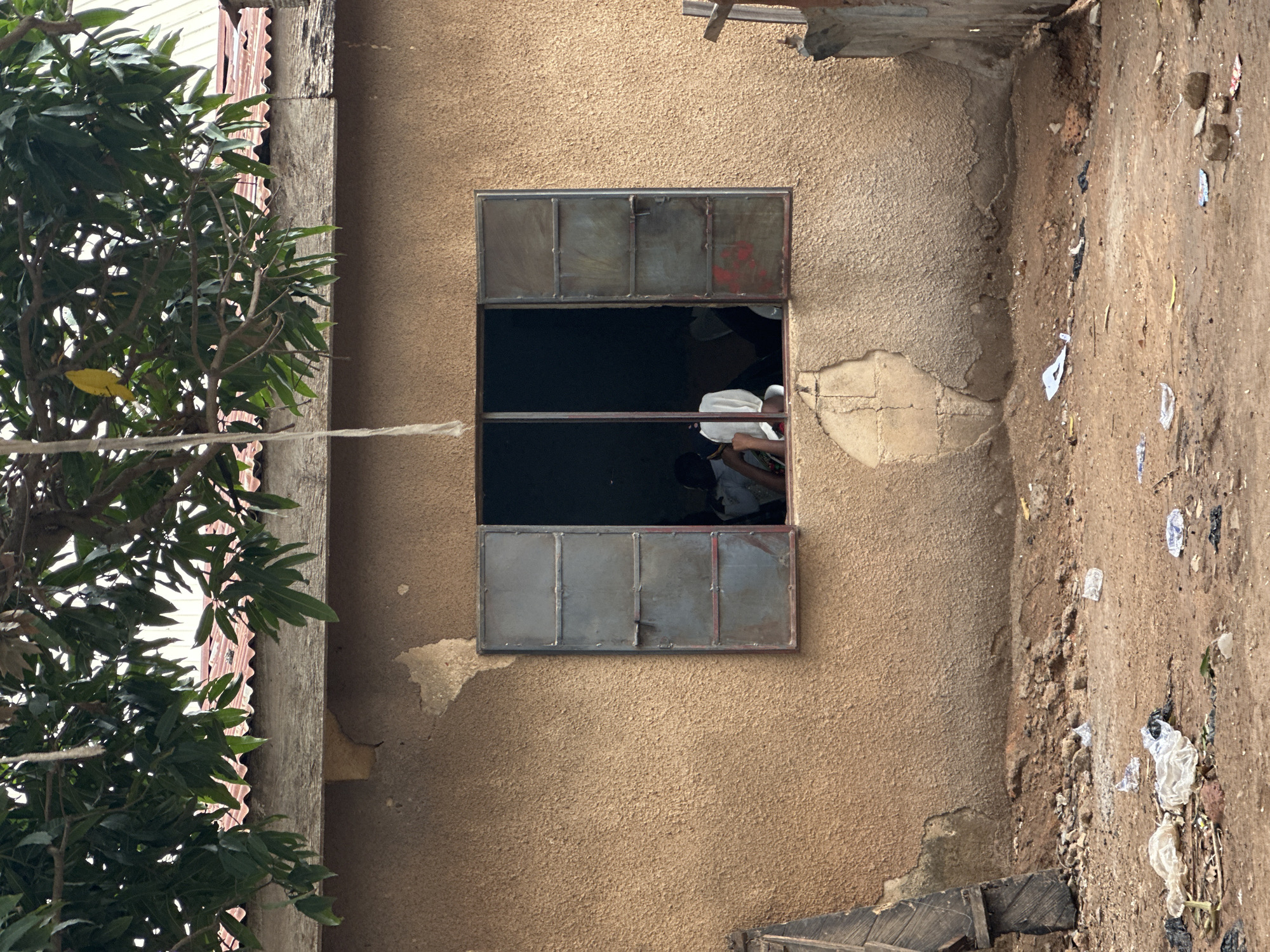 An exterior forensic image depicts a section of a single-story building in an unknown, unknown city location, viewed from an unpaved outdoor area. The building features a rough, textured, light-brown stucco wall showing patches of wear and exposed sub-layers. A rectangular window with an aged metal frame is centered in the wall; its two outward-opening shutters, painted a faded blue-grey with red accents, are both fully open. Inside the dimly lit room, several individuals are partially visible: one person wearing a white garment or head covering is prominent on the left, with others' silhouettes seen to their left and right. Above the window, a weathered wooden fascia board supports a reddish-brown corrugated metal roof, largely obscured by the dense green foliage of an overhead tree. A thin, light-colored rope hangs vertically from a tree branch, and a single yellow leaf is visible among the green leaves. The foreground consists of uneven, reddish-brown dirt, liberally scattered with various items of refuse, including multiple clear plastic bags, white paper, and other debris. On the far left, a dark, weathered wooden plank leans against the building. On the far right, a small wooden post protrudes from the ground.