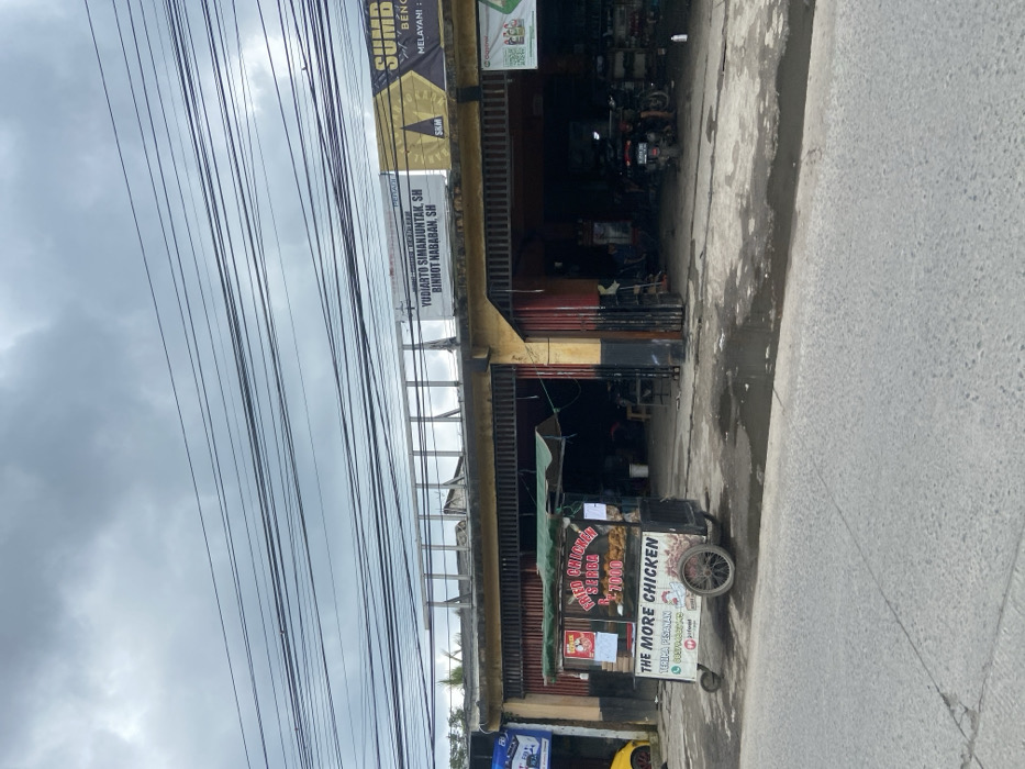The image shows a street scene in Baleendah, Indonesia. There is a food cart parked on the left side of the street, with a sign that reads "Fried Chicken Rp. 7.000". The cart is in front of a yellow building with a red door. On the right side of the street, there is a large building with a sign in Indonesian, which appears to be an advertisement. Several power lines run across the scene. There are a few motorbikes parked in front of the building, and a person is walking towards the camera. The street is wet and has puddles of water. The sky is grey and cloudy.