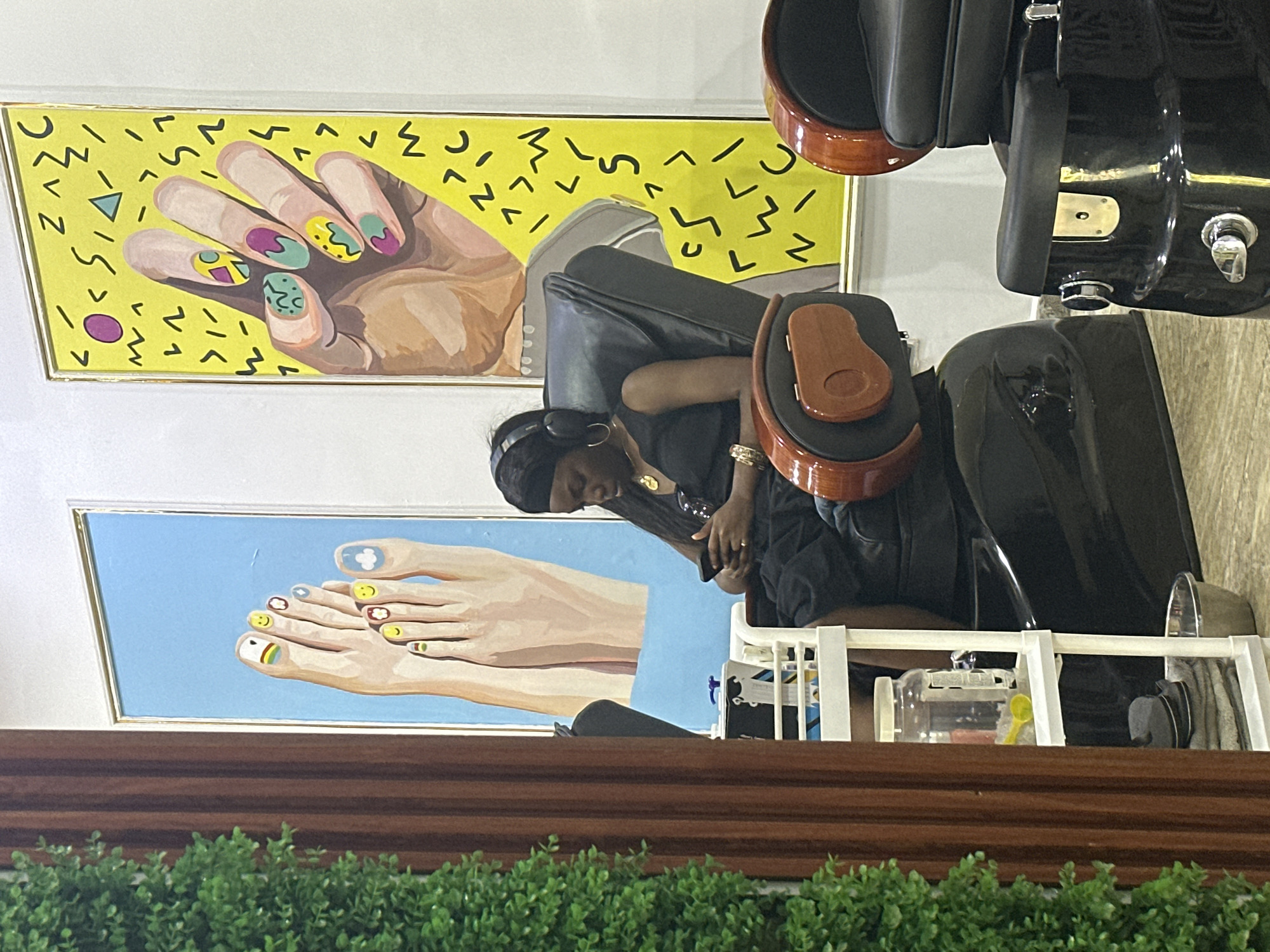 This image depicts an indoor scene, likely a nail salon, with two large, colorful paintings on the wall. The painting on the left is of a pair of feet with brightly colored nail polish on each toe, including designs like a rainbow, smiley faces, and a cloud. The painting on the right is of a hand with similarly decorated fingernails, set against a yellow background with abstract shapes.
In the foreground, a woman is seated in a black, reclined salon chair. She is wearing headphones and looking down at her phone, which she is holding in her hands. She has a black headscarf on and is wearing a black top. She is also wearing jewelry, including a necklace and bracelets. To her right, another identical salon chair is partially visible.
To the left of the woman and her chair, there is a white shelving unit with various items, including a large clear jar filled with liquid, a yellow scoop, and some folded gray towels. A metallic basin is also visible on the floor near the shelving unit. The left edge of the frame shows a tall, vertical structure covered in artificial green foliage, possibly a decorative plant or screen. The overall environment appears to be clean and well-lit, suggesting a commercial establishment. The context of Abuja, Nigeria, places this scene within a specific urban setting, though no explicit indicators of this location are visible within the frame itself, beyond the possibility of the style and decor being indicative of the region.
