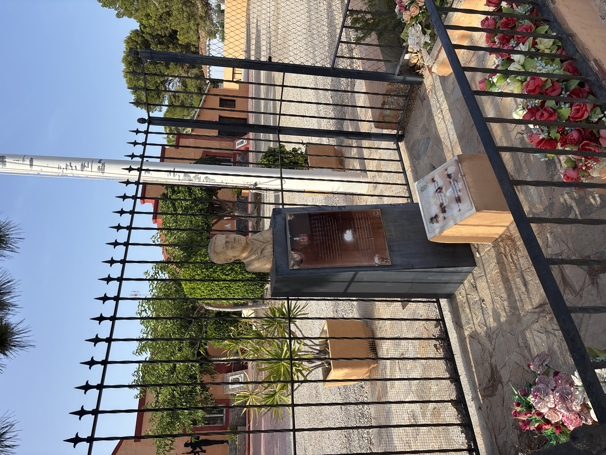 The image depicts an outdoor monument in Orihuela, Spain, likely dedicated to a military figure. In the foreground, a black metal fence encloses a paved area with decorative flowers. Behind the fence, a statue of a man's bust sits atop a dark stone pedestal. In front of the pedestal, a smaller plaque is mounted on a light-colored, stepped structure. The background reveals a building with orange walls and a fence, along with trees and a clear blue sky. The overall scene suggests a memorial site or a public space with historical significance.