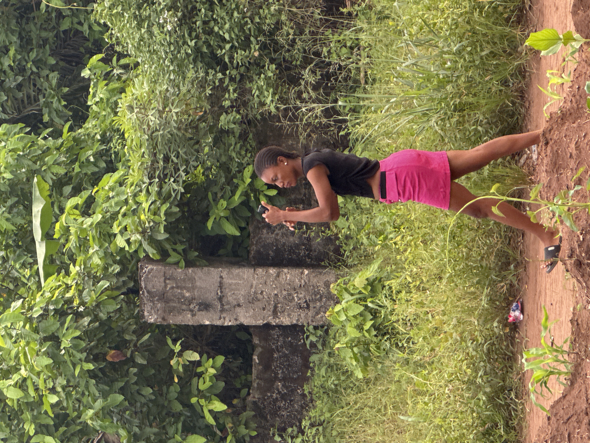 A medium, outdoor shot depicts a woman in Aboh, Nigeria, looking at a handheld device. She is wearing a black shirt and pink skirt. She stands on a dirt path next to a field of green grass and foliage. To her left is a weathered concrete structure surrounded by leafy plants.