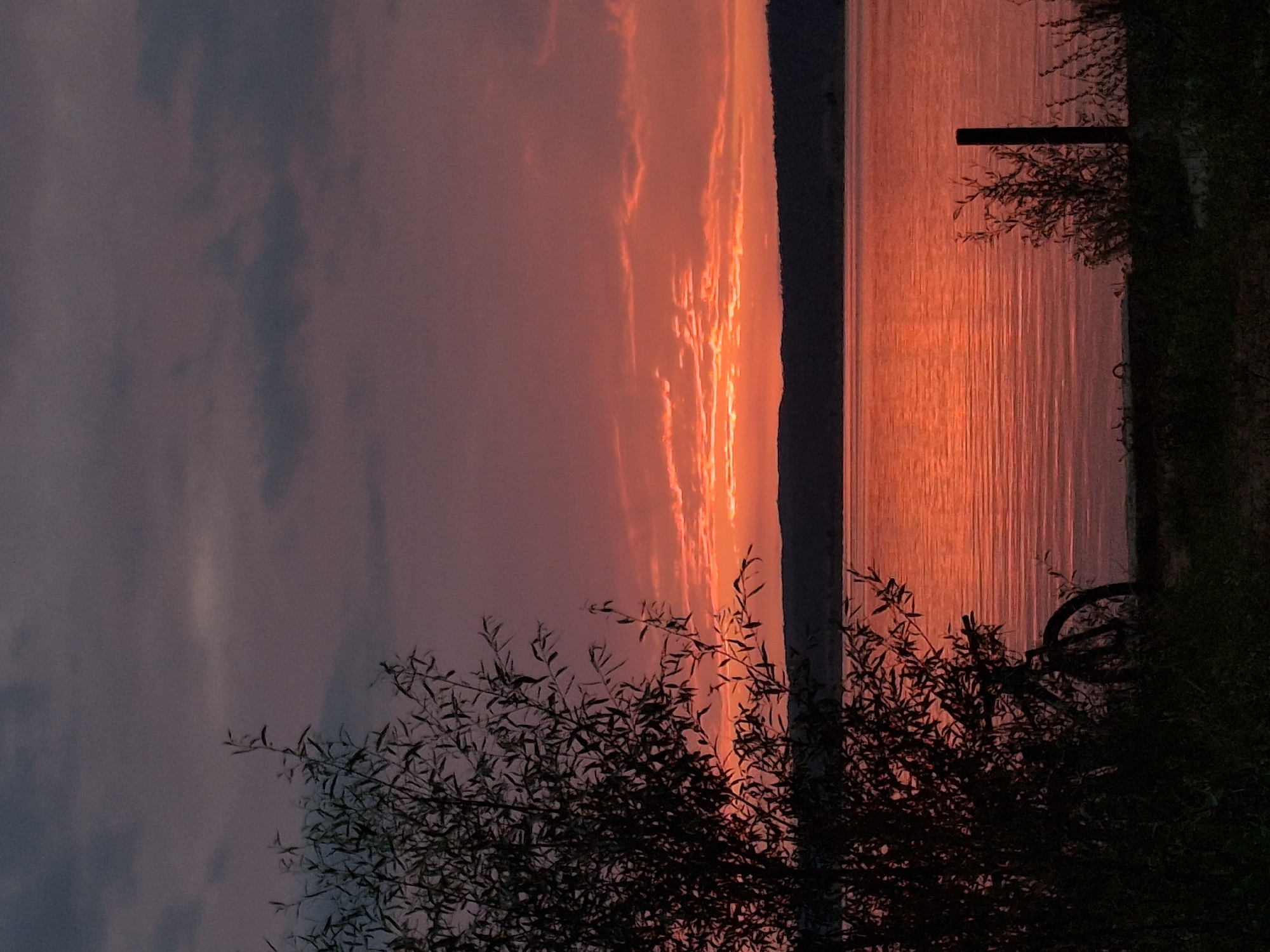 A serene sunset over the Kama River in Chaikovsky, Russia. The sky is a dramatic canvas of deep purples and vibrant oranges, with wispy clouds catching the last rays of sunlight. The river reflects the fiery colors of the sky, creating a shimmering, almost liquid gold surface. In the foreground, silhouetted against the twilight, is a lone bicycle parked near a cluster of trees and a fence post. The scene is peaceful and still, evoking a sense of quiet contemplation.