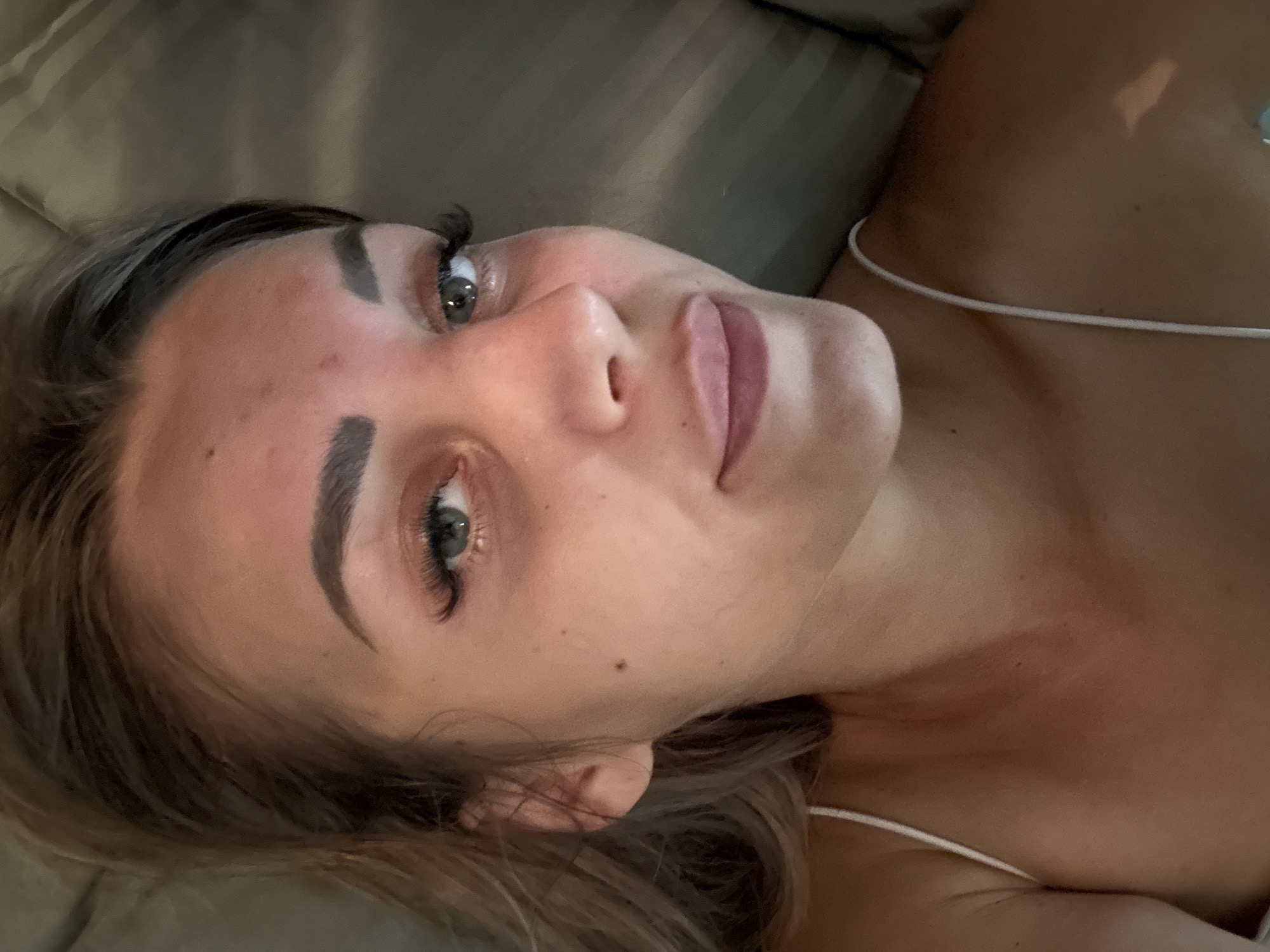 The image shows a close-up, overhead shot of a young Caucasian woman lying down. She is looking towards the camera with a neutral expression. Her hair is blonde and pulled back. She is wearing a white spaghetti strap top. The background appears to be a bed with grey linens. There are no identifiable landmarks or context within the image to indicate a specific location in Yuzhno-Sakhalinsk, Russia.