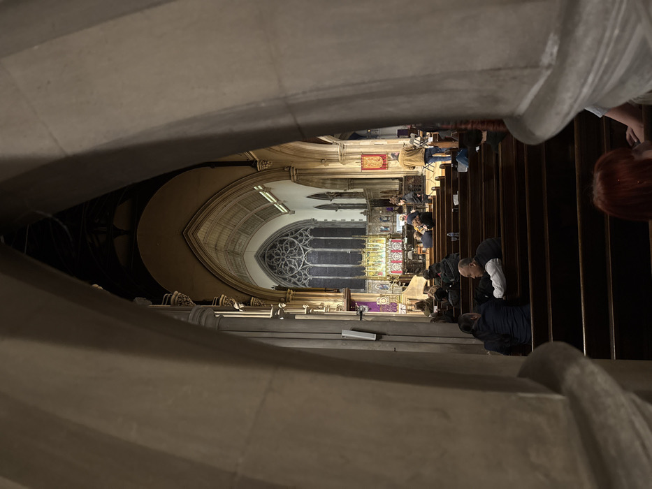 The image depicts the interior of St George's Church, Bloomsbury, in the city of London, United Kingdom. The perspective is from the nave, looking towards the chancel and altar, framed by two large, light-colored stone arch supports in the immediate foreground.
The environment is a large, dimly lit Victorian Gothic Revival church. The nave is furnished with rows of dark wooden pews. Numerous individuals are seated in these pews, facing the chancel. Their attire is varied, generally casual. In the lower left-midground, two men are seated, one in a white shirt and dark vest, the other in a dark jacket. A person with reddish-brown hair is partially visible in the bottom right.
In the mid-distance, the chancel area is visible, brightly lit compared to the nave. A group of musicians is present, positioned to the right of the altar. One individual, wearing a light brown jacket, stands near a music stand, appearing to play an instrument or conduct. Other musicians are seated.
The chancel features an elaborate, multi-tiered reredos behind the altar, characterized by numerous niches, possibly statuary, and prominent gilded elements. Above the reredos, a large Gothic East Window displays intricate stone tracery, including a distinctive large central circular motif in its upper portion and multiple vertical lights below. Purple liturgical banners are draped on the left side of the chancel, hanging from stone architectural features. A white speaker unit is mounted on a pillar on the left side of the nave. Several water bottles are visible on the ledges of the pews. The overall scene suggests a musical performance or rehearsal within a church setting during an evening or low-light period.