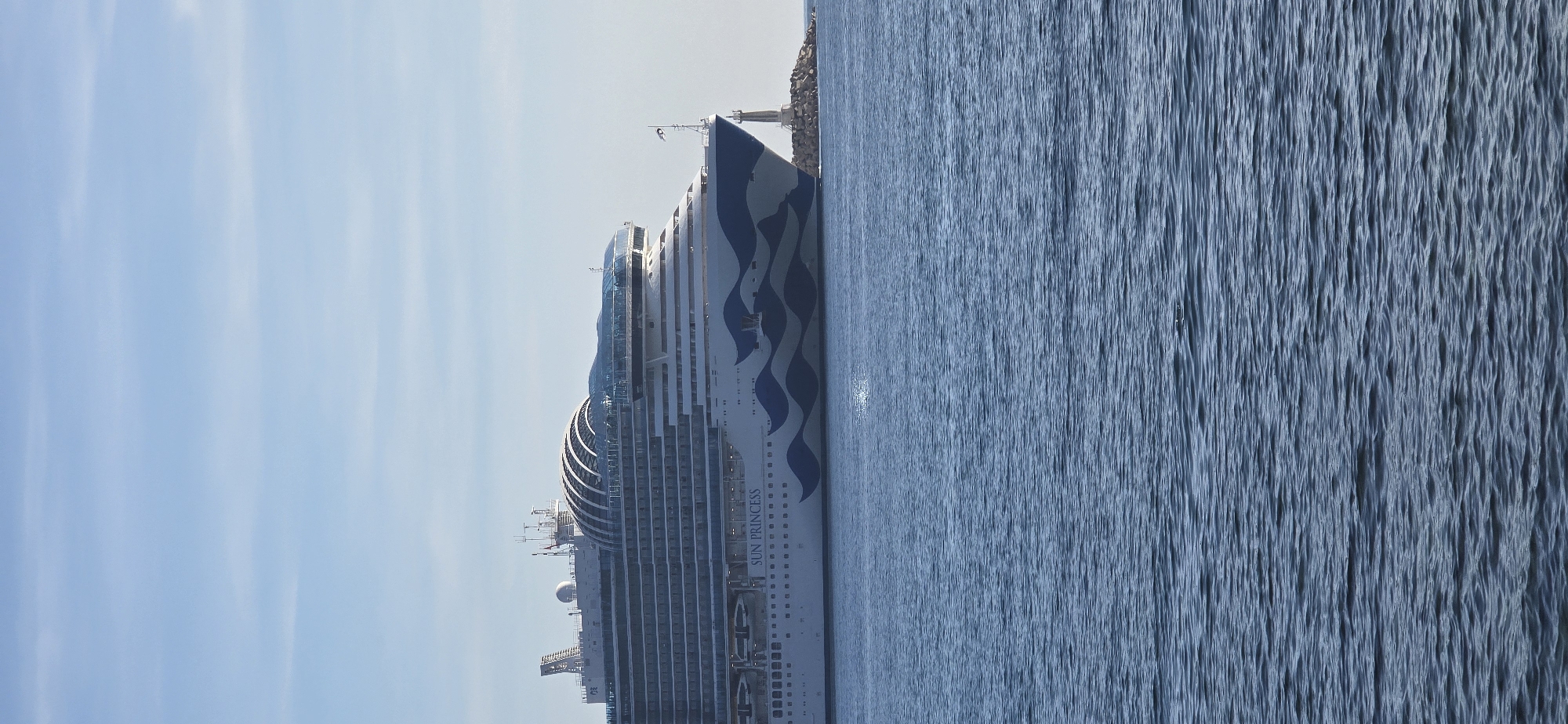 This daytime photograph captures the cruise ship "SUN PRINCESS" in the waters near Bar, Montenegro. The vessel, clearly identified by its name printed on the hull, features a white body with a striking blue, wave-like pattern. Multiple decks, balconies, and a distinctive glass-domed structure on its upper levels are visible.
The ship is positioned on the left side of the frame, facing towards the right. The foreground and right side are dominated by the calm, blue-grey sea, which exhibits numerous ripples and a bright glint of sunlight reflecting on its surface. Beyond the ship's bow, a rocky breakwater or coastline is visible, topped with a small, white structure resembling a lighthouse or beacon. The sky above is a clear, light blue, with only faint, wispy cloud formations.
The scene suggests calm weather conditions, and the ship appears to be stationary, possibly docked or anchored in the coastal area. No people are visible.