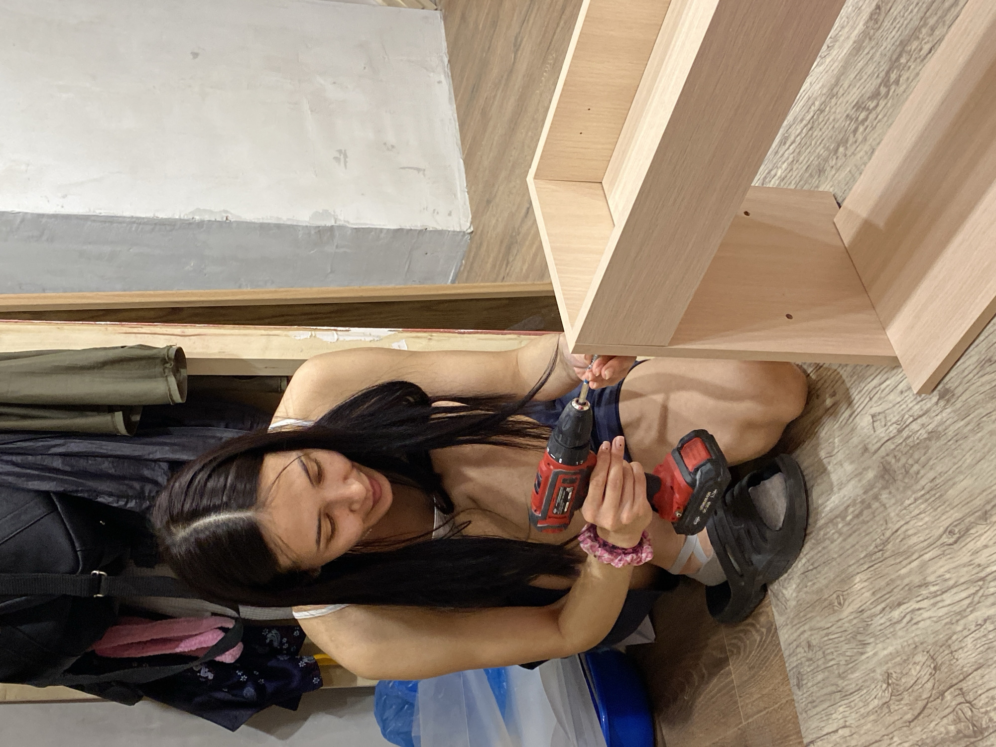 A woman is kneeling on a wood-patterned floor, using a red and black drill to assemble a light-colored wooden structure that appears to be part of a shelving unit. She is fair-skinned, has long black hair, and is wearing dark clothing. A pink scrunchie is on her wrist. To her left is a collection of items including a black backpack with a pink object, a blue plastic bag, and some dark clothing. Behind her, against the wall, is a rough, unfinished surface with a visible seam. The room seems to be under construction or renovation. The location in Votkinsk, Russia cannot be determined from the given image.