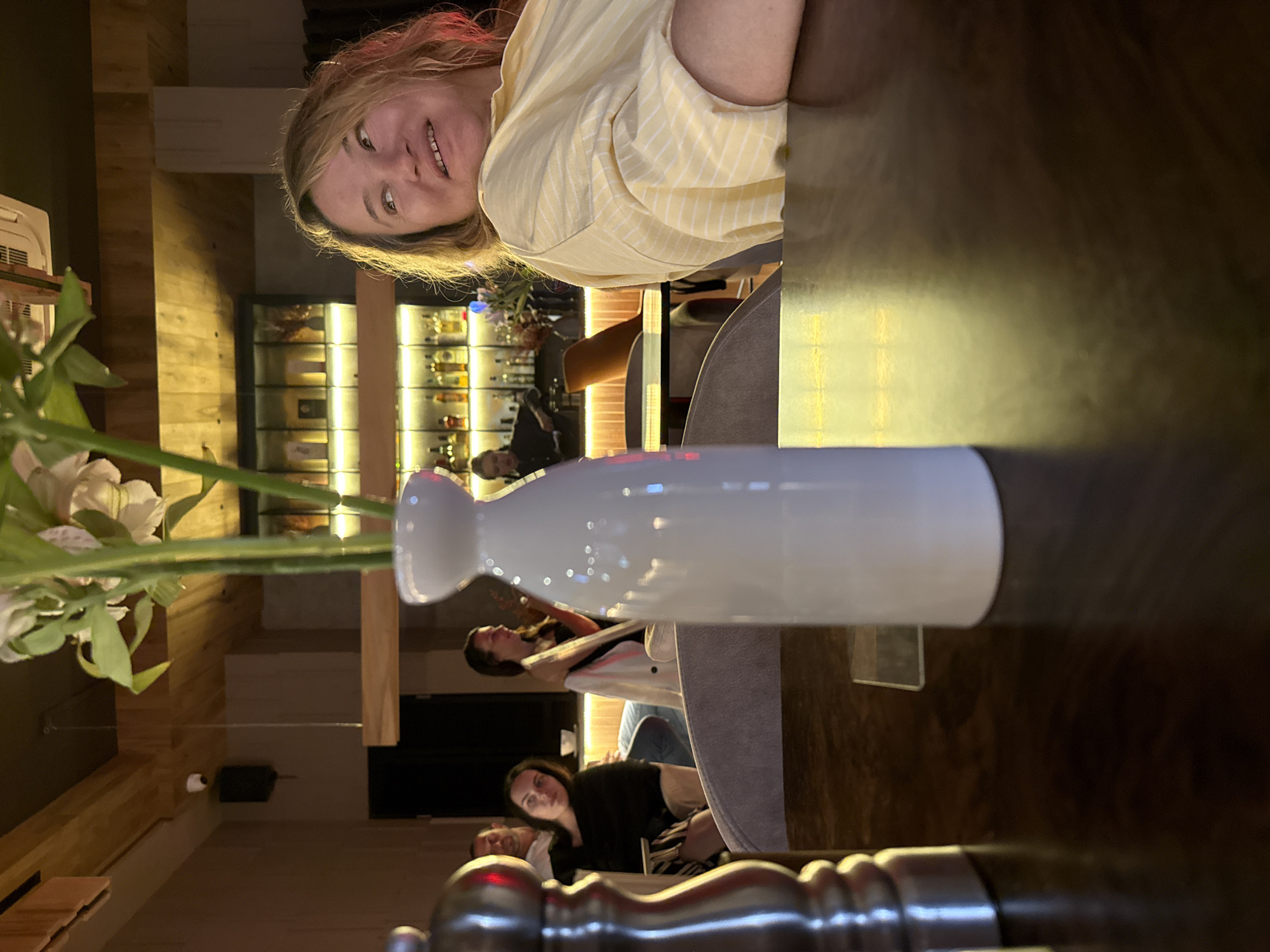This is a portrait-oriented, somewhat blurry photo taken indoors, likely at a restaurant or bar. The primary subject is a woman with blonde hair, smiling and looking towards the camera. She is wearing a light yellow, striped shirt. In the foreground, a white ceramic sake bottle and a vase with white flowers are visible, partially obscuring the view of the background.
Behind the woman and the sake bottle, the setting appears to be a dimly lit establishment with wooden accents. A bar area is visible in the background, with shelves stocked with bottles and illuminated by warm lighting. There are other patrons in the background, some seated and some standing, though their features are indistinct. The overall atmosphere seems relaxed and social. The photo appears to have been taken at night due to the interior lighting and dark surroundings. The location context of Sochi, Russia is noted, but there are no specific visual cues within the image to confirm this. No visible text is present in the image.