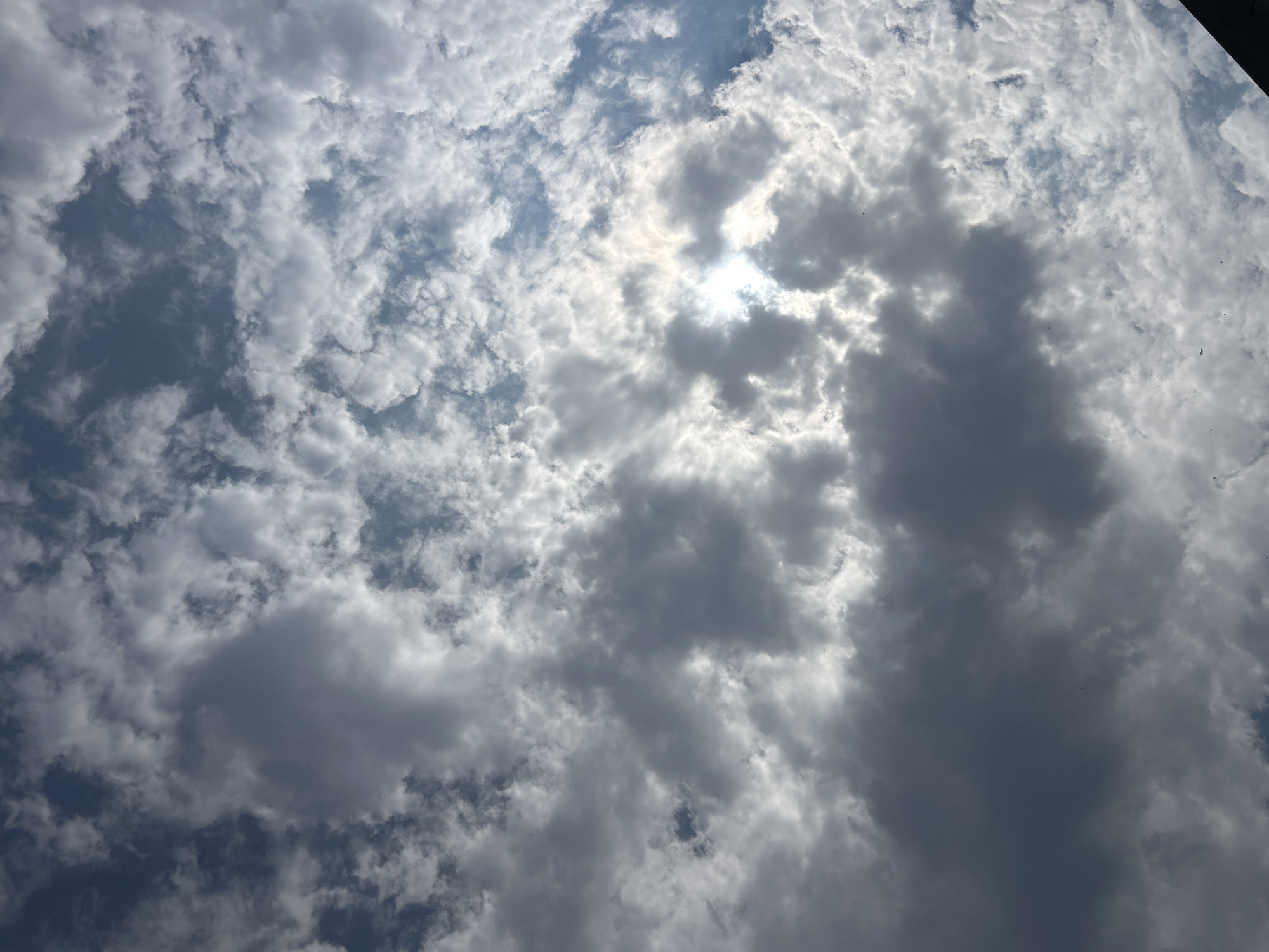 A low-angle, full-frame shot captures a cloudy sky above Giussano, Italy. The sun, partially obscured by dark, cumulus clouds, creates a bright, hazy glow in the upper right quadrant of the image. Scattered, wispy white clouds fill the rest of the frame, interspersed with patches of blue sky. The composition is entirely focused on the atmospheric conditions, with no discernible man-made structures or living beings visible. The overall impression is one of an overcast day with intermittent sunshine.