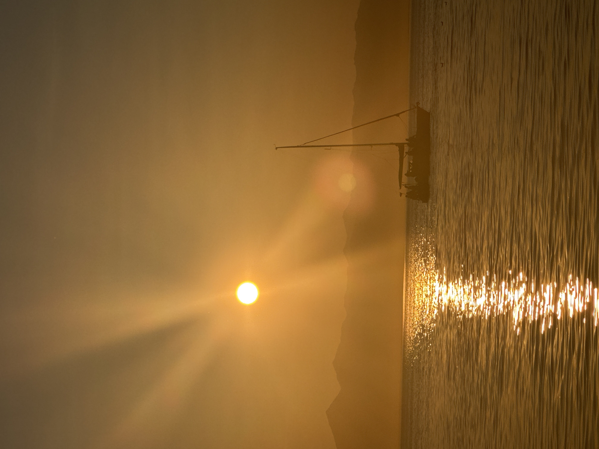 The image depicts a serene seascape at sunset or sunrise, bathed in warm golden light. The sun, a bright, luminous orb, is positioned in the upper left quadrant of the frame, casting long, dramatic rays across the sky. In the foreground, the calm water reflects the golden hues of the sky, creating a shimmering path of light. A sailboat, silhouetted against the vibrant backdrop, is the primary subject. The boat appears to be moving, with several figures visible on its deck, suggesting a leisurely voyage. In the distance, a range of mountains forms a hazy, muted silhouette against the horizon. The overall atmosphere is one of tranquility and natural beauty. The image does not contain any identifiable landmarks or features that would specifically place it in the city of Petropavlovsk-Kamchatsky, Russia.