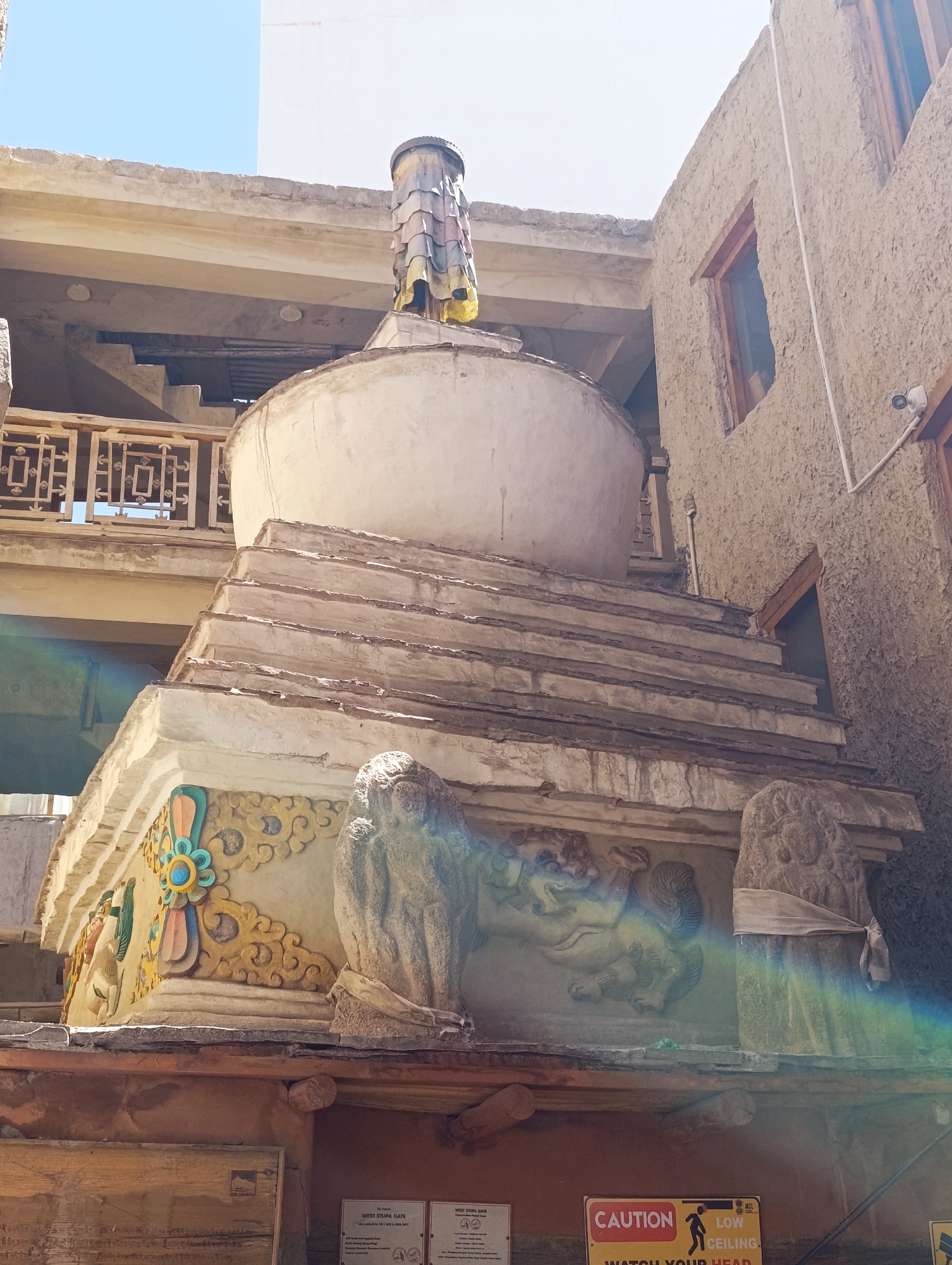 A Buddhist stupa, adorned with intricate carvings of lions and floral motifs, stands prominently in the old city of Leh, India. The stupa's base features a series of tiered levels, with the main body being a large, rounded dome. The structure is made of stone and plaster, showing signs of age and weathering. Decorative elements include colorful painted designs on the lower section of the stupa, and carved lion figures flanking the entrance. Above the dome, a cylindrical structure with layered, colorful panels rises towards the sky, topped by what appears to be a tire.
Surrounding the stupa are buildings constructed with traditional Ladakhi architecture, featuring rough, textured walls and wooden window frames. To the left, a balcony with an ornate railing overlooks the scene. The sky above is a clear, bright blue, indicating a sunny day. A faint rainbow-like lens flare is visible in the lower right portion of the image, suggesting the presence of strong sunlight. In the foreground, a wooden beam supports the structure, and signage indicating "CAUTION LOW CEILING" is visible, warning visitors of a potential hazard. No people are visible in the frame, but the presence of the signage suggests it's a public area, likely a tourist site within Leh.