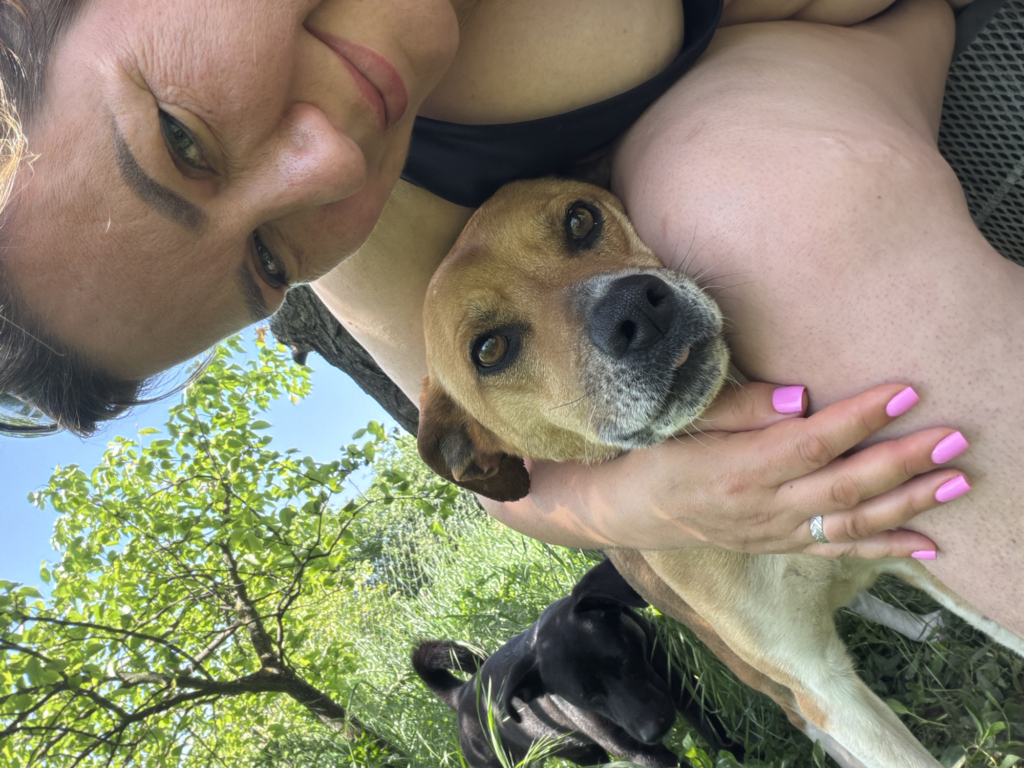 A selfie shot depicts a woman holding a tan dog close to her body. A second, black dog is visible in the background among green grass and foliage. The location is a grassy outdoor area, likely in Leningradskaya, Russia. The woman has fair skin and pink nail polish. The tan dog looks directly at the camera.