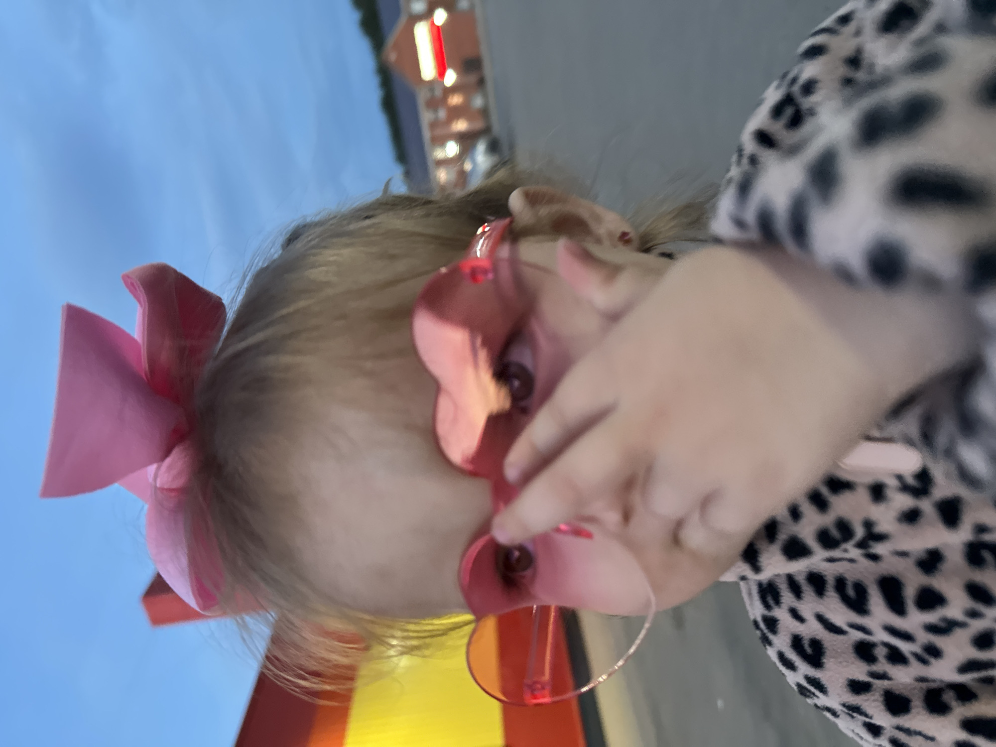 A close-up, slightly blurry image shows a young child wearing large pink heart-shaped sunglasses and a pink bow in their hair. The child is wearing a leopard-print jacket and is partially obscuring their face with their hand. In the background, there are buildings with illuminated signs and a parking lot with a few cars. The sky is a dusky blue, suggesting it is either early morning or late evening. The location is Chaikovsky, Russia.