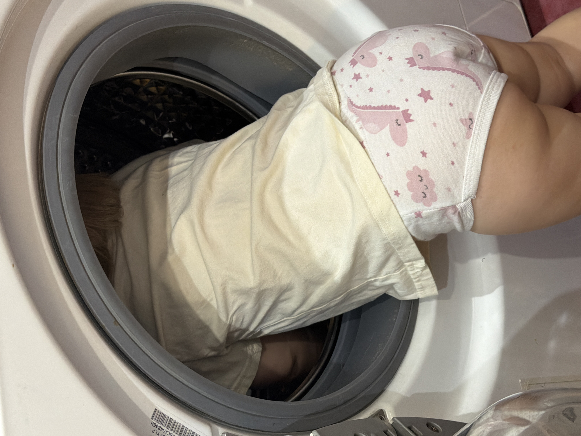 A child wearing a light yellow shirt and underwear with pink dinosaur and star patterns is partially inside the drum of a front-loading washing machine. The child's head is inside the drum, and their lower body is outside the machine. The washing machine is white, and the surrounding environment is not clearly visible. The location is identified as Chaikovsky, Russia.
