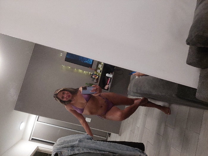 A light-skinned female individual, standing barefoot and wearing a two-piece lilac bikini, is depicted in a mirror selfie within an indoor setting. She holds a light purple smartphone in her right hand, angled towards the mirror, and smiles directly into the lens. Her left arm is slightly bent, with her hand on her hip, and she poses with her right leg crossed in front of her left. The reflection reveals a modern apartment interior with light gray tiled flooring and predominantly gray walls. To the left, a denim jacket hangs on a hanger next to a dark gray sliding-door closet. In the background, a wall-mounted dark gray shelf unit displays various bottles and a white cylindrical object. Above the shelf, a flat-screen television shows a blue and purple illuminated image, beneath which warm-toned string lights are draped. A section of a dark gray upholstered couch is visible in the lower right foreground. The specific location within Buenos Aires, Argentina, cannot be determined from the visual information provided in the image.