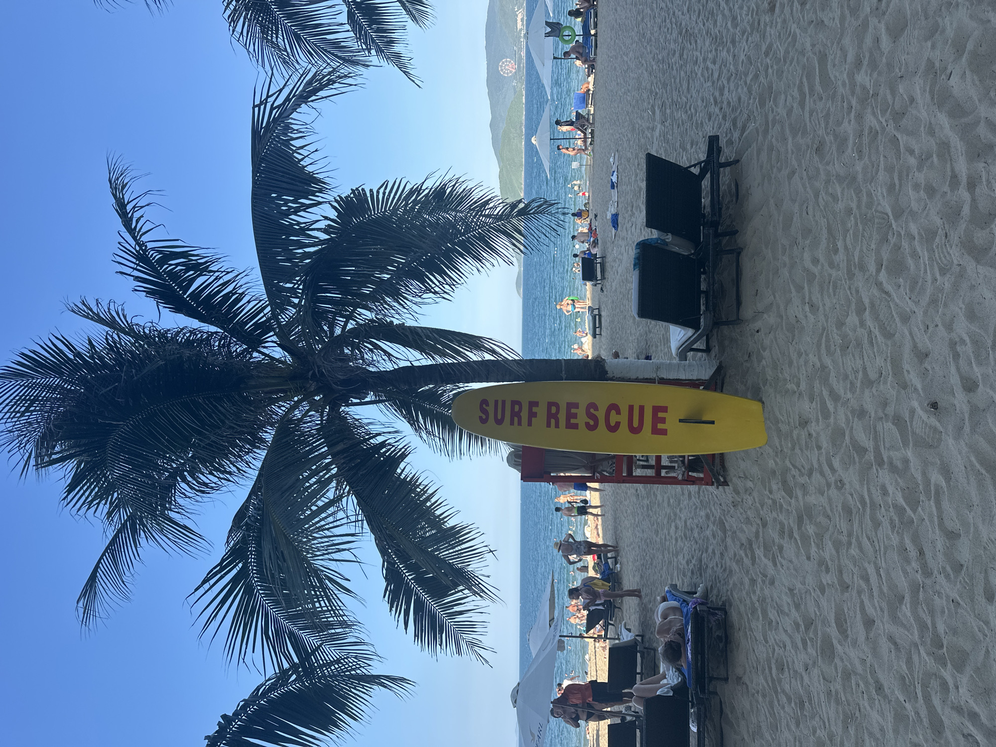 A bright, sunny day at a beach in Nha Trang, Vietnam, under a clear blue sky. The foreground is dominated by fine, light-colored sand, with a few dark beach chairs and a yellow surfboard propped against a red lifeguard stand. The surfboard is emblazoned with the words "SURF RESCUE" in red lettering.
In the mid-ground, the sandy beach stretches out, populated by numerous sunbathers on lounge chairs and towels, as well as people walking and wading in the shallow waters of the ocean. Several white umbrellas provide shade for beachgoers. The ocean itself is a vibrant blue, with gentle waves lapping at the shore.
The background features a hazy coastline with green hills and a Ferris wheel visible in the distance. Palm trees frame the scene, their fronds casting shadows on the sand and adding to the tropical ambiance. The overall impression is one of a lively and relaxed beach resort.