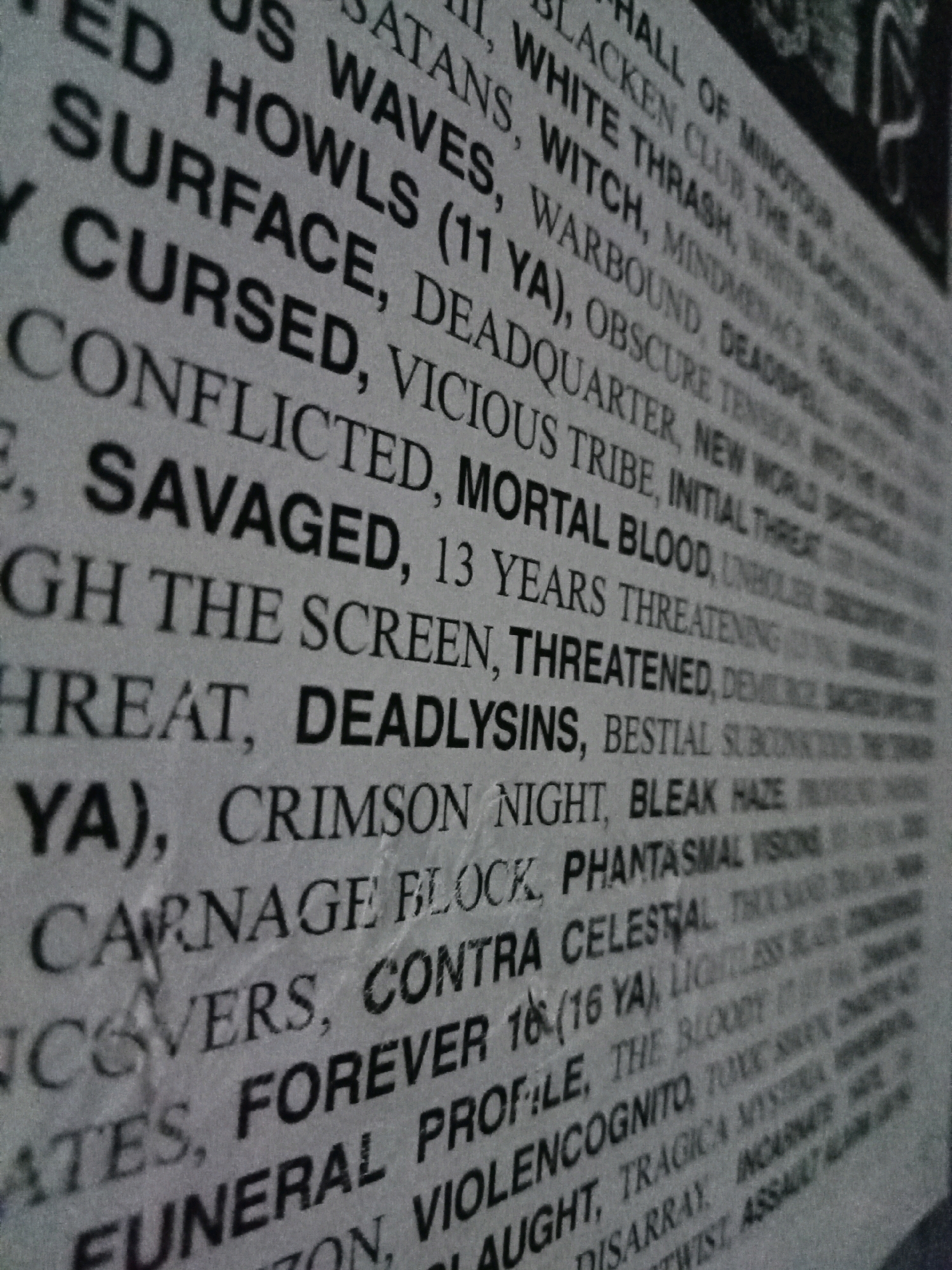 The image is a close-up, angled shot of a poster with small, black text printed on a gray, textured background. The text appears to be a list of words or phrases, possibly song titles, band names, or descriptive terms, arranged in diagonal lines. Some words are capitalized and appear to be emphasized, such as "SAVAGED", "THREATENED", and "DEADLYSINS". The printing is slightly blurred and the texture of the paper is visible, suggesting the image may have been taken with a mobile phone or a camera with a shallow depth of field. There are no people or specific landmarks visible in the image. The image does not contain any information that would allow for identification of its location within Depok, Indonesia.