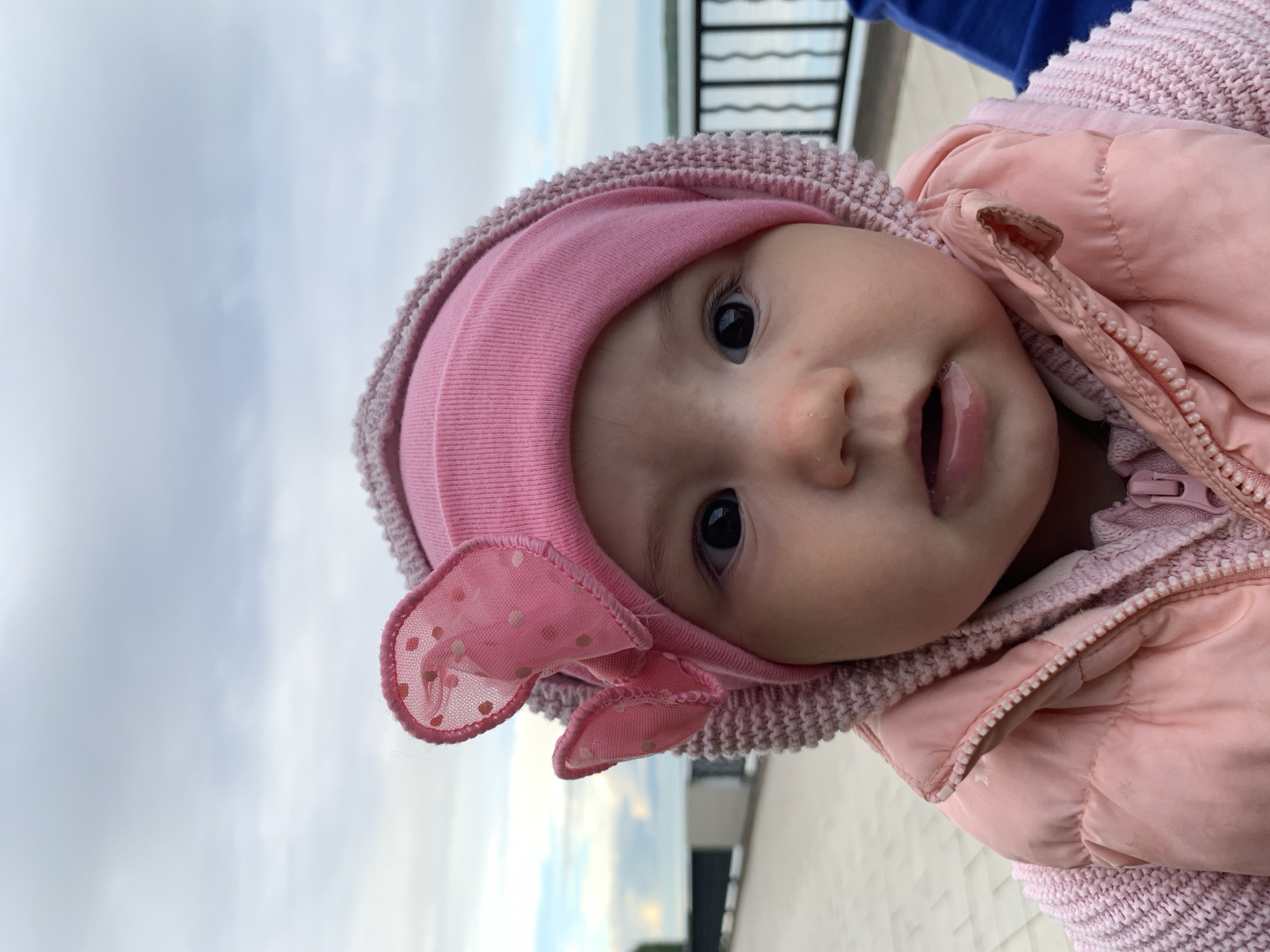A close-up, tilted shot shows a baby wearing a pink knit hat with a pink polka-dot bow and a pink quilted jacket. The baby is looking at the camera with an open mouth. The background shows a cloudy sky, a body of water, and a railing. The image was taken outdoors in daylight. It is not possible to determine if the image was taken in Chaikovsky, Russia, or to determine specific actions or interactions from the image.