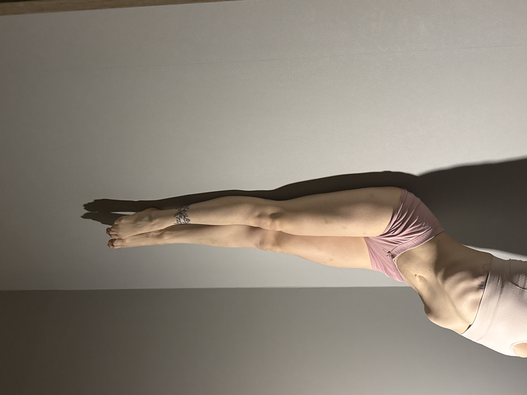 This image captures a slender individual, likely female, positioned horizontally against a plain, light gray background, indicative of an indoor setting. The person is wearing light pink athletic-style underwear or shorts and a matching sports bra or crop top. Their body is posed with legs extended and torso slightly arched, showcasing a very lean physique with ribs visibly defined. A decorative tattoo, appearing as an abstract or leafy band, is visible around the left ankle.
Strong, directional lighting creates prominent, elongated shadows of the person's body on the gray surface, suggesting a single light source from the side or above. The upper portion of the background appears brighter, gradually darkening towards the bottom. The individual's face is not visible, making emotional assessment impossible, but the pose appears controlled and deliberate, possibly for a photograph or as part of a static exercise.
The scene lacks specific location cues within the frame, but the context indicates the image was captured in Chaikovsky, Russia. There is no visible text in the image.