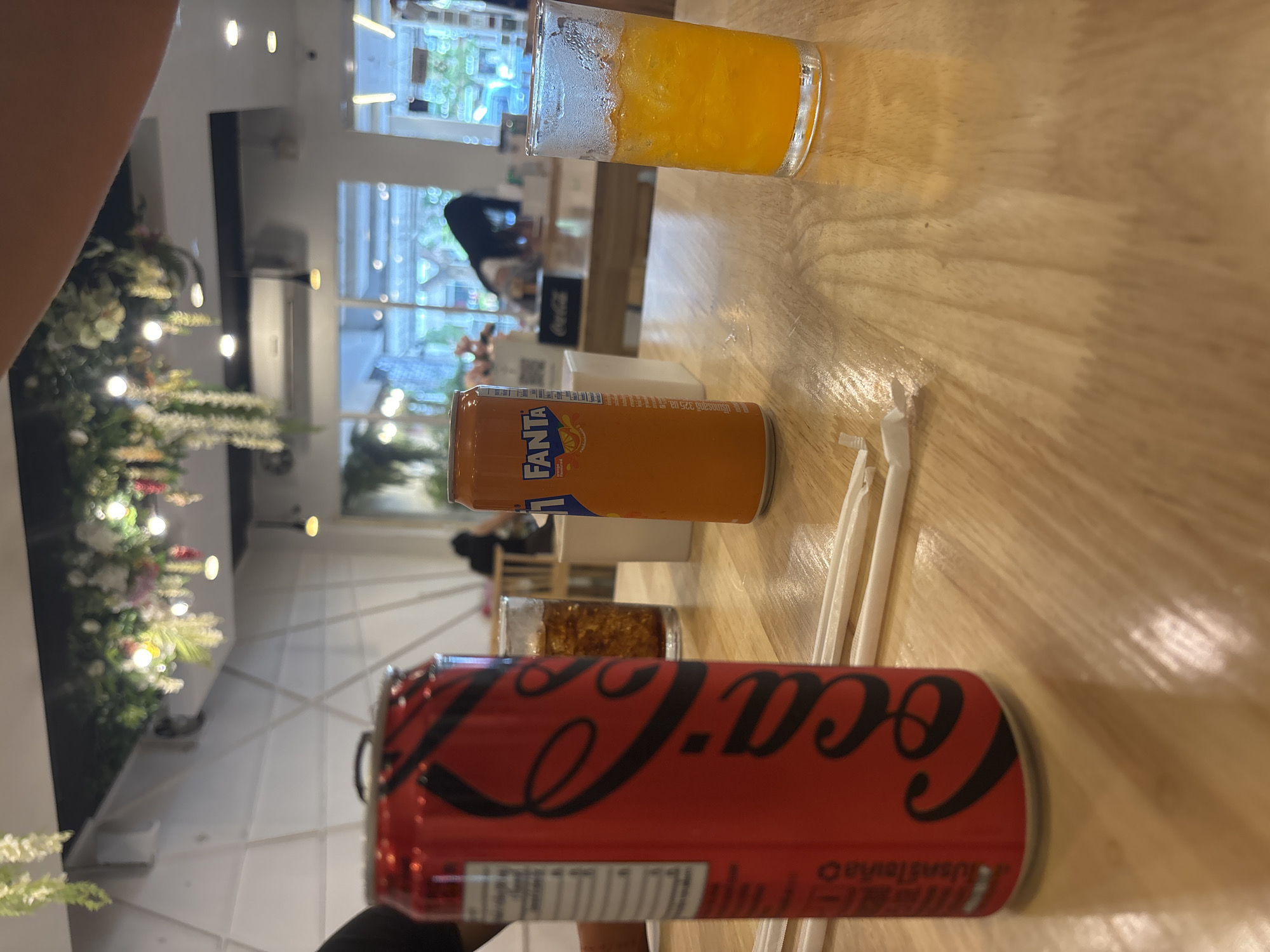 A low-angle, medium shot captures a wooden table in what appears to be a restaurant setting in Chiang Mai City Municipality, Thailand. In the foreground, a red Coca-Cola can is prominently displayed on the left, with its iconic white lettering visible. To its right, an orange Fanta can stands next to a glass filled with a vibrant orange liquid, likely Fanta or orange juice, with ice cubes. Several white paper straws are scattered on the table, suggesting that drinks have been prepared or are about to be consumed. In the background, the restaurant's interior is blurred, showing glimpses of other tables, chairs, and customers. The ceiling is adorned with hanging floral decorations and soft lighting, creating a pleasant ambiance. The overall scene depicts a casual dining experience with refreshing beverages.