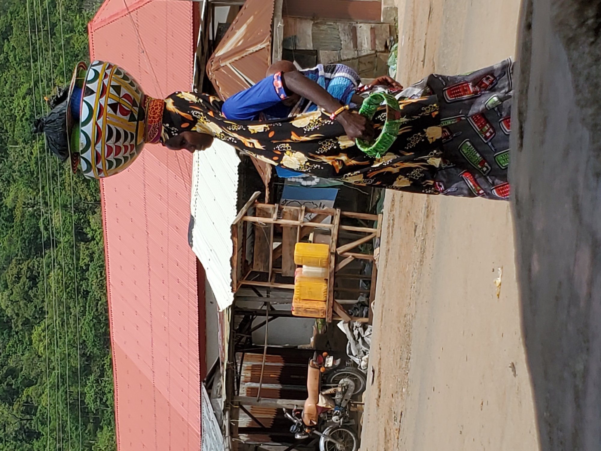 A woman walks down a street in Mararaba, Nigeria, carrying a large, decorated bowl on her head. She wears a patterned dress, and another person in blue clothing is nearby. A motorcycle is parked under a makeshift wooden structure. The area appears to be a commercial or residential street with buildings on either side, one with a red roof and a partial view of forested land in the background. The street surface is dirt or gravel.